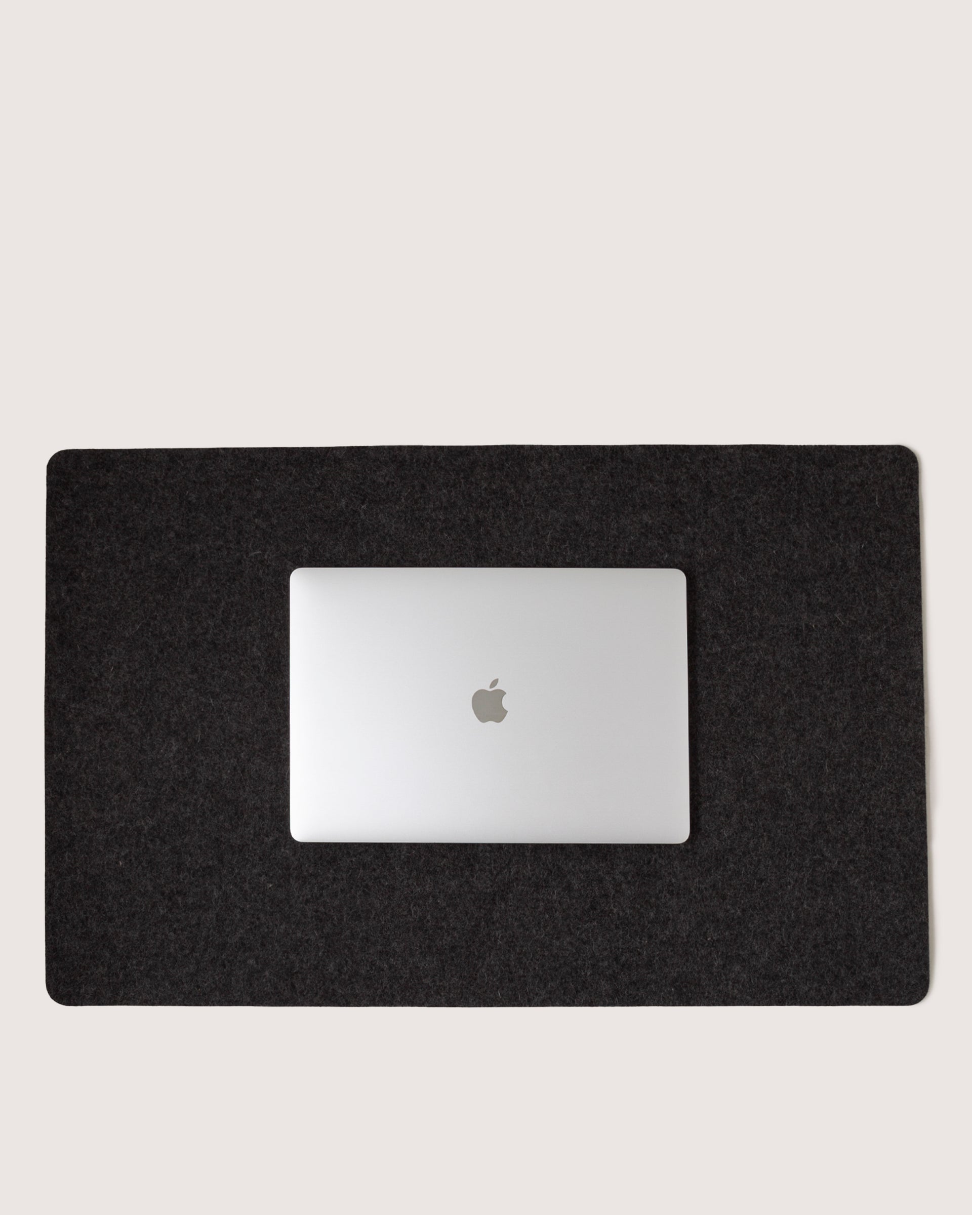 A closed silver laptop with an Apple logo sits on a graf lantz Mosen Desk Pad made of Merino Wool, centered on a large black rectangular mat against a plain light background.