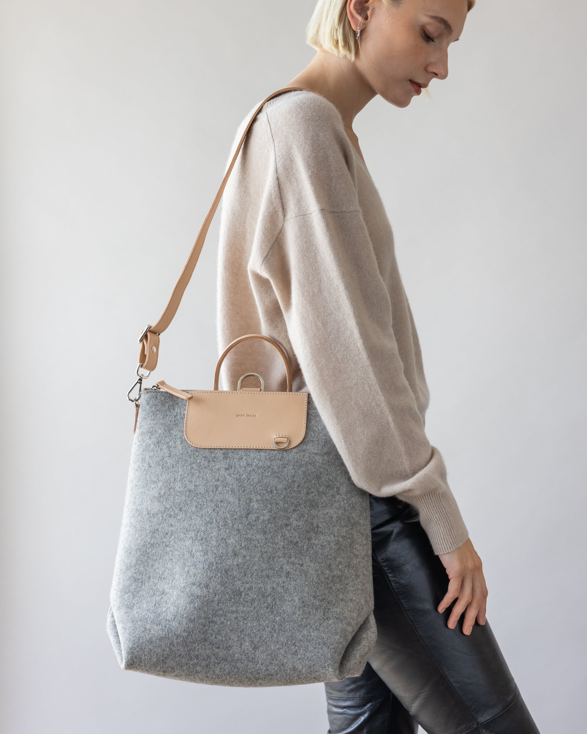 A person with short blonde hair is wearing a beige sweater, black leather pants, and holding a stylish graf lantz light grey and beige handbag made from sustainable material against a plain white background.