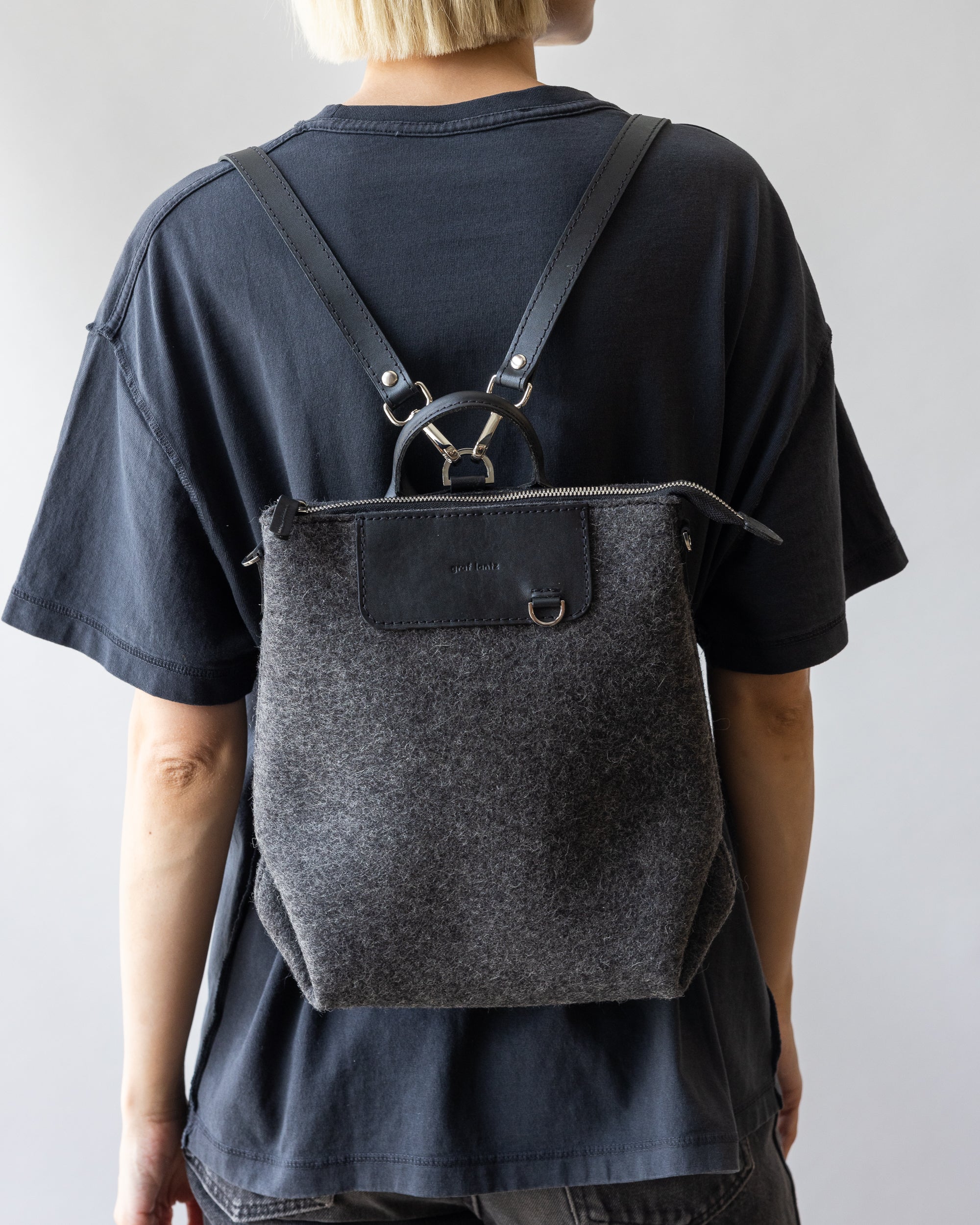 A person with short blonde hair carries a graphite gray felt backpack from graflantz, made of sustainable materials and featuring versatile black leather straps. They wear a loose-fitting black T-shirt and pants, standing against a plain light gray background.