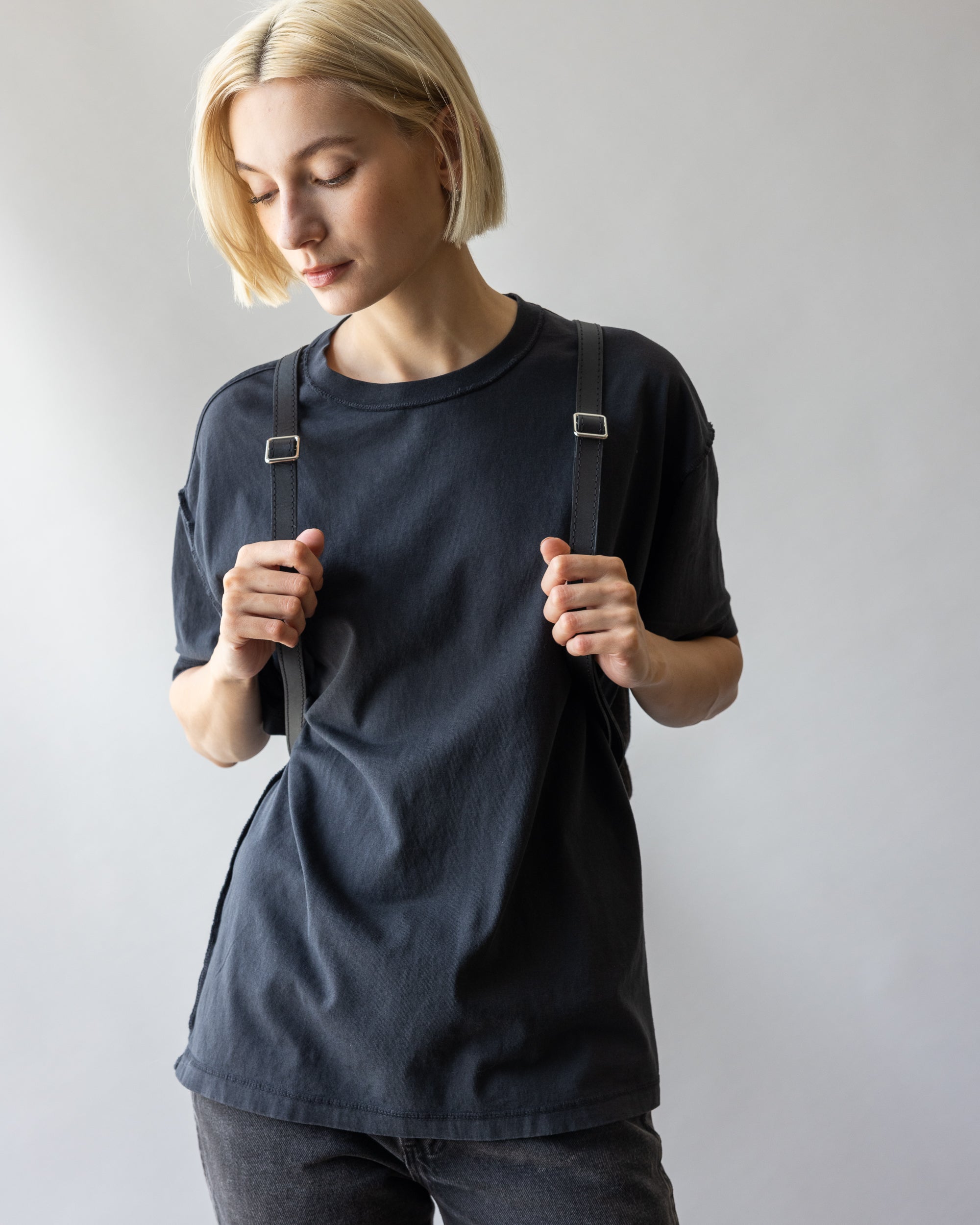 A person with short blonde hair in a black T-shirt holds the straps of a graf lantz versatile bag against a softly lit gray background.
