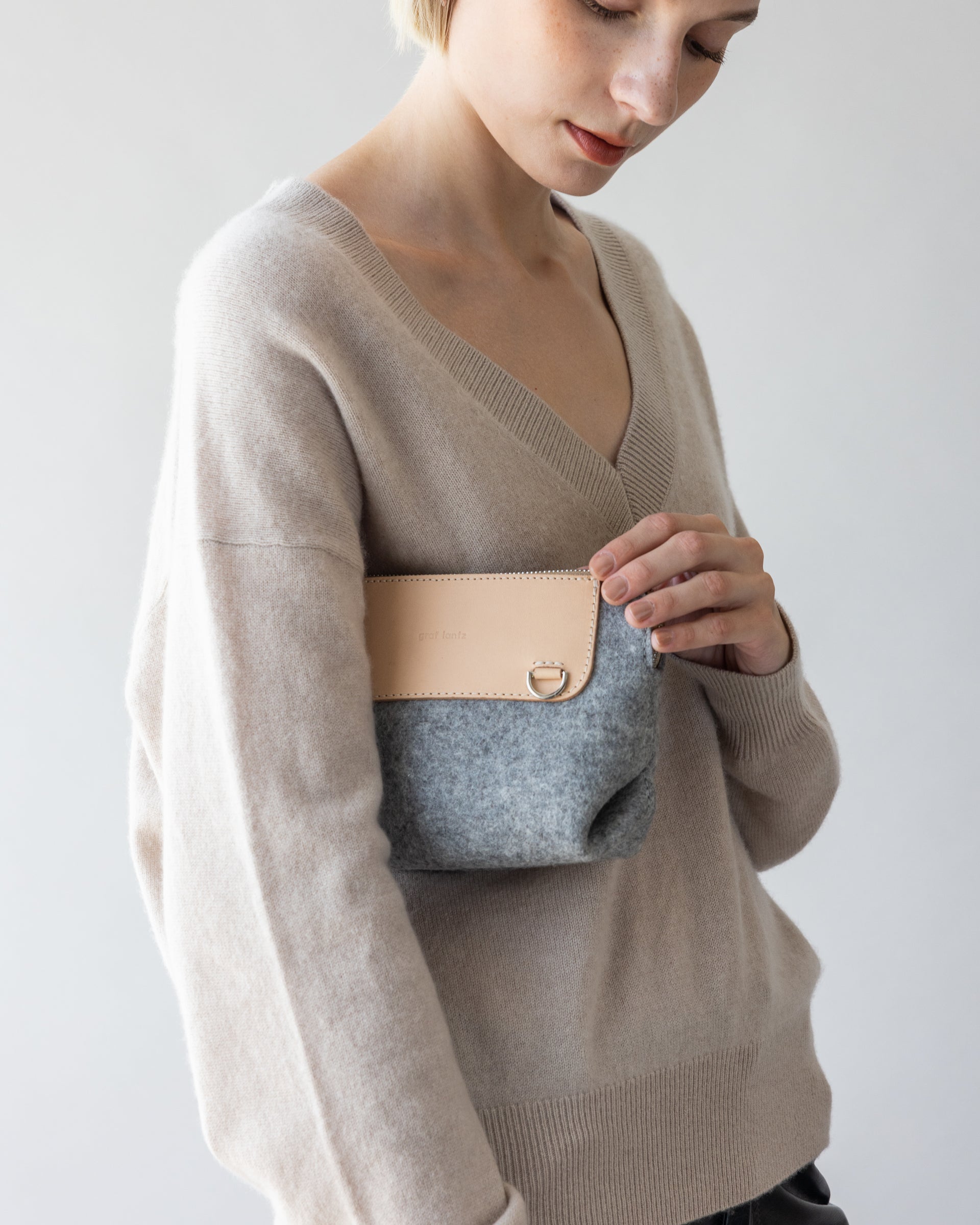 A person wears a beige sweater of soft merino wool and clutches a small graf lantz bag with a gray body and tan flap, exuding cozy elegance against a light background while hinting at hands-free convenience.