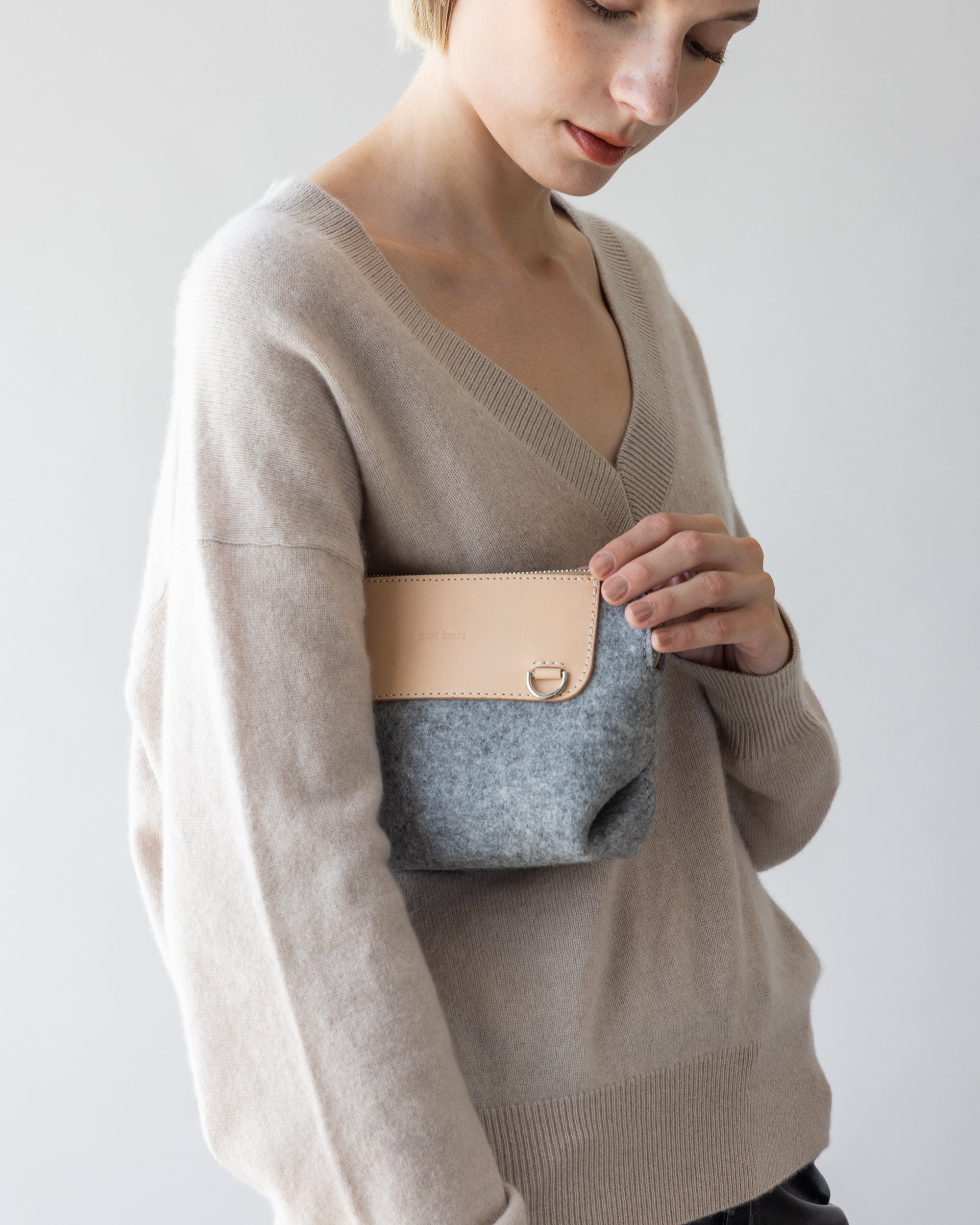 A person wears a beige sweater of soft merino wool and clutches a small graf lantz bag with a gray body and tan flap, exuding cozy elegance against a light background while hinting at hands-free convenience.