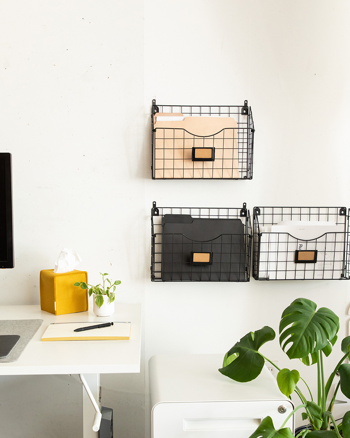 A minimalist workspace includes a white desk with a tissue box and plant. Against the wall, graf lantz wire baskets hold file folders in peach, black, and vegetable tan leather. A green plant is nearby, partially hiding the edge of a computer monitor.