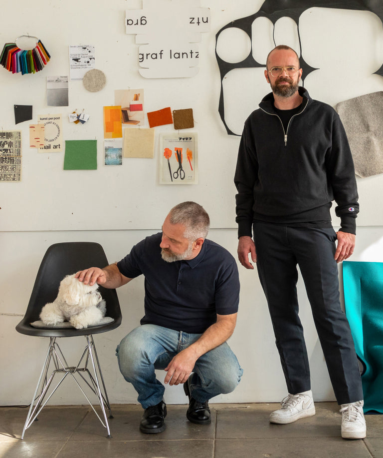 Daniel Lantz kneels next to white dog Frankie on a black chair next to standing Holger Graf, founders of Graf Lantz