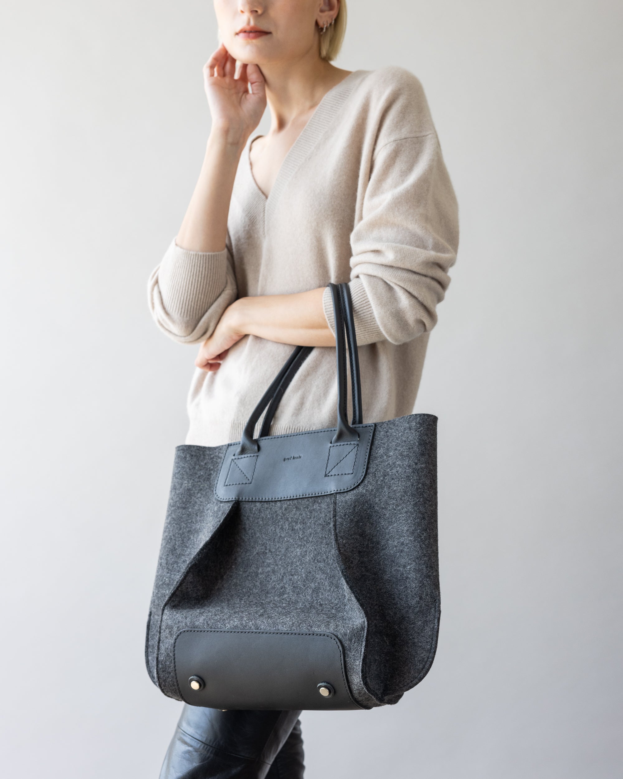 A person wearing a beige Merino wool sweater and dark pants holds a large gray tote bag featuring black leather accents and magnetic closure. The plain, neutral background accentuates the minimalist design of this Frankie Midi bag by graf lantz.