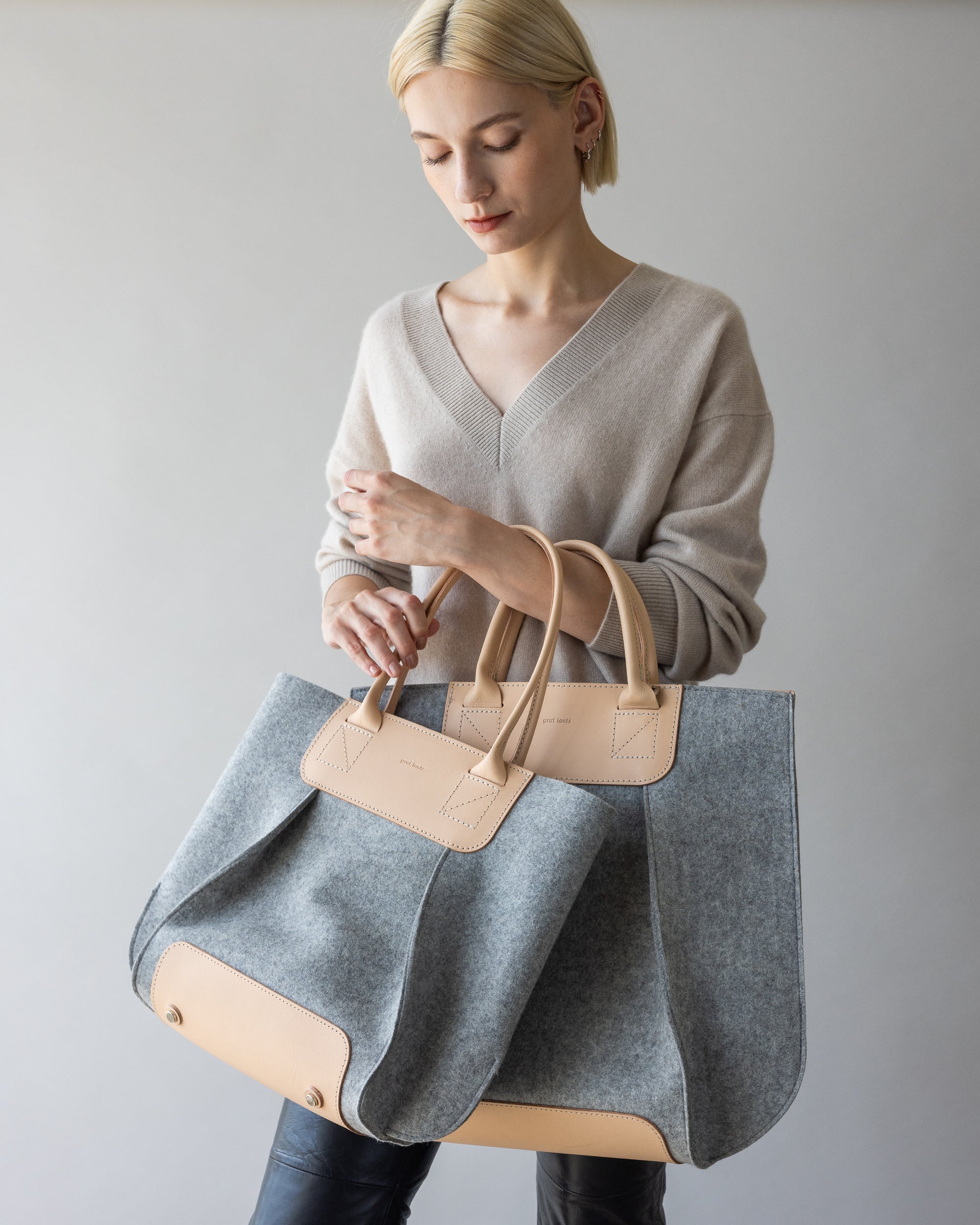 A short-haired blonde person, dressed in a beige sweater and black pants, holds a graf lantz large gray felt tote with tan leather accents. Against a light gray backdrop, they embody the chic vibe of Frankie Midi effortlessly.