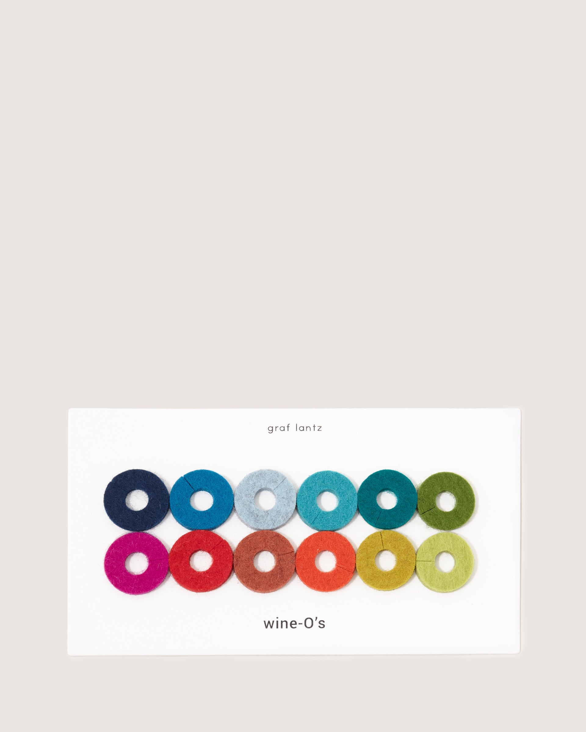 A white card displays 12 colorful wine-Os felt rings in two rows, made from Bavarian Merino wool by graf lantz. Each vibrant ring marks a wine glass in shades of blue, green, red, and pink.