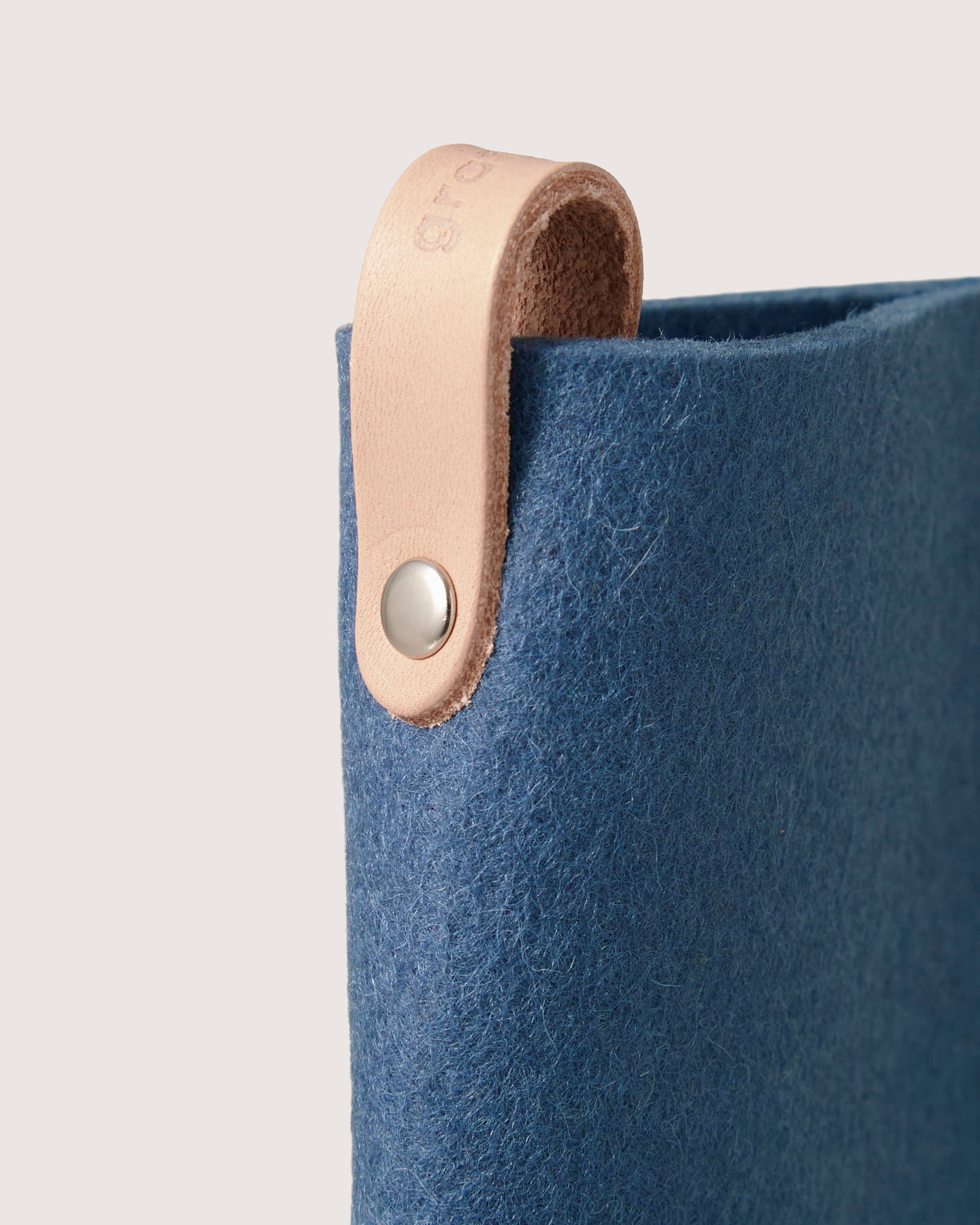 A close-up of the graf lantz blue Merino wool felt with a beige leather strap, ideal for crafting an elegant eyeglass sleeve. The metal button fastens the strap into a loop at the top against an off-white background that highlights its texture and color beautifully.