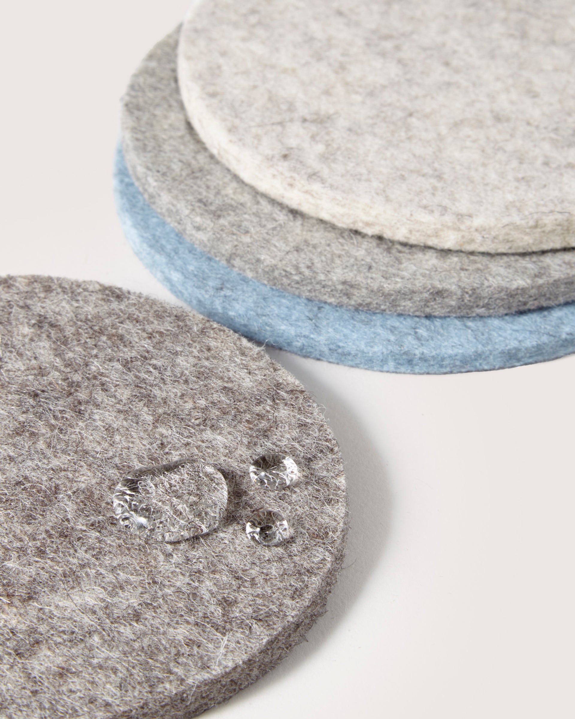 A stack of graf lantz round felt coasters in gray, blue, and cream rests on a light surface. Made from German Merino wool, the top coaster sparkles with droplets, showcasing its soft, fuzzy texture.