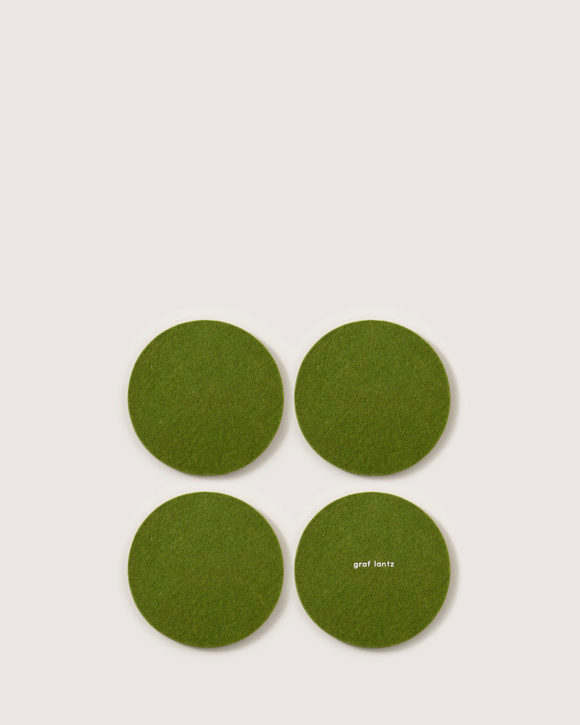 Four green felt coasters, made from German Merino wool and shaped round, are placed in a square on a light background. The coaster at the bottom right prominently features the brand name graf lantz.
