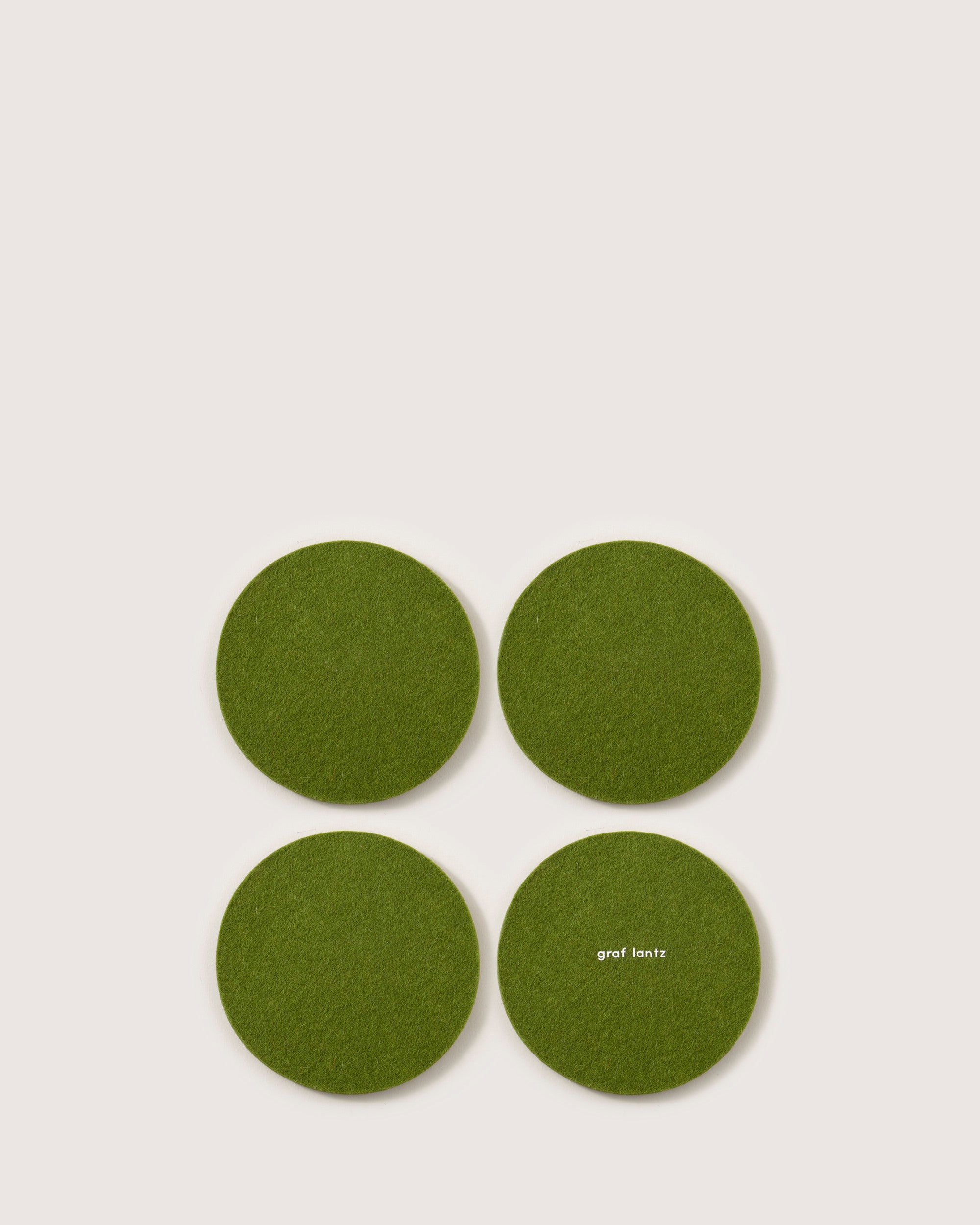 Four green felt coasters, made from German Merino wool and shaped round, are placed in a square on a light background. The coaster at the bottom right prominently features the brand name graf lantz.