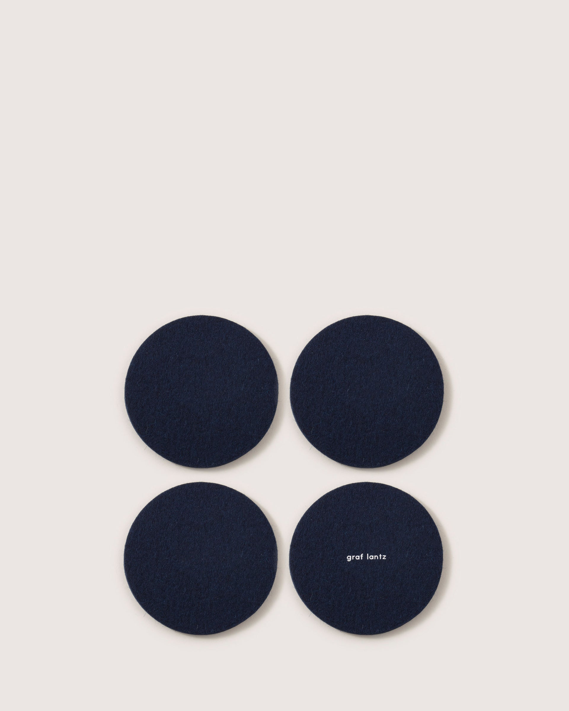 Four dark blue circular coasters made from premium Merino wool are arranged in a square on a light background, with one displaying the graf lantz brand name.