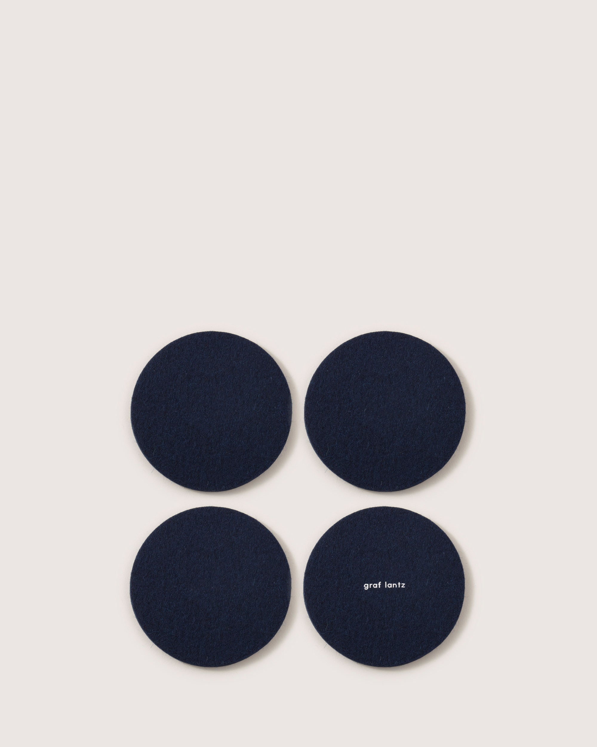 Four dark blue circular coasters made from premium Merino wool are arranged in a square on a light background, with one displaying the graf lantz brand name.