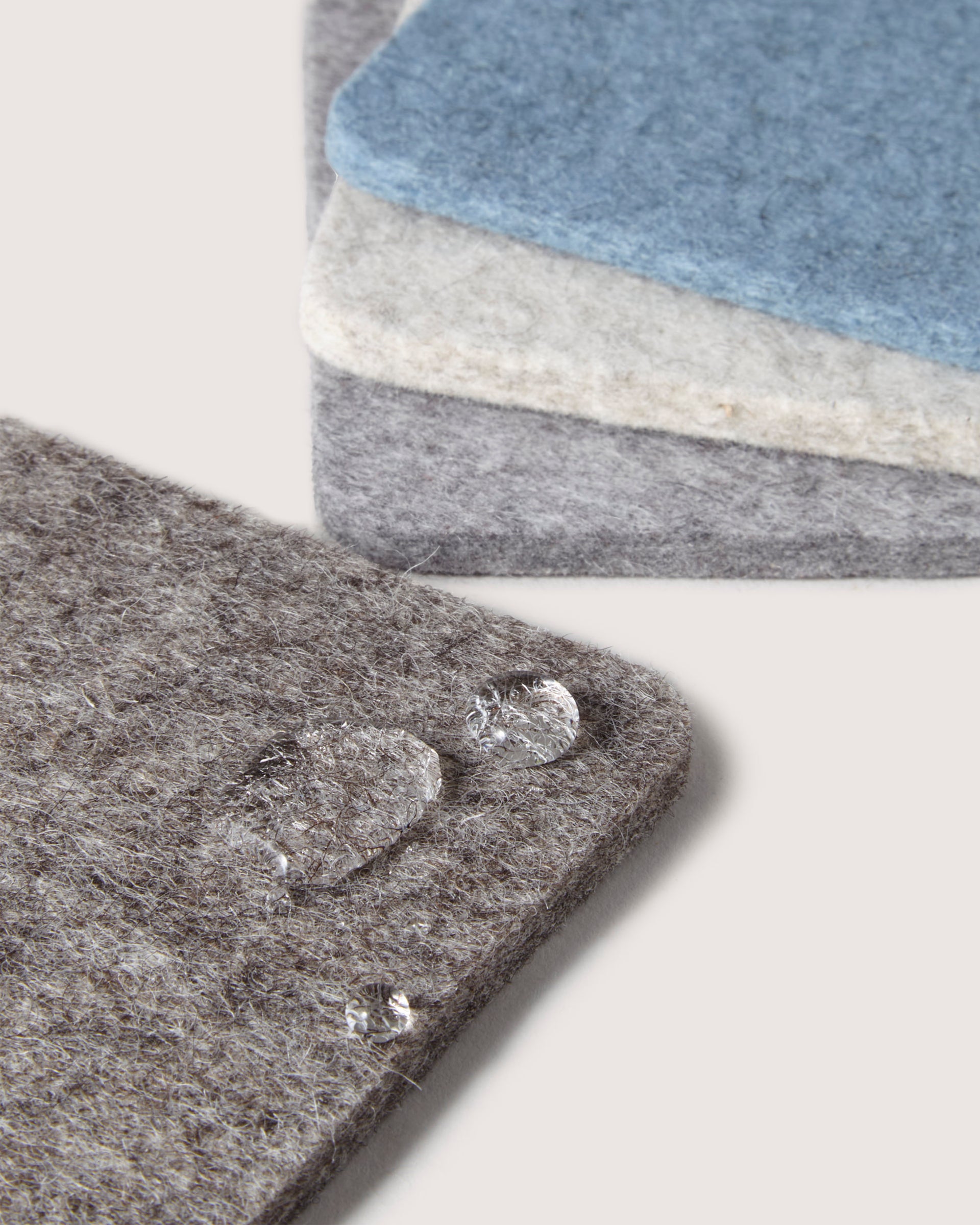 Close-up of three graf lantz felt coasters made from Merino wool in gray and blue hues, with the gray one on top. Water droplets highlight their texture and water-resistant quality, adding stylish appeal to any home décor.