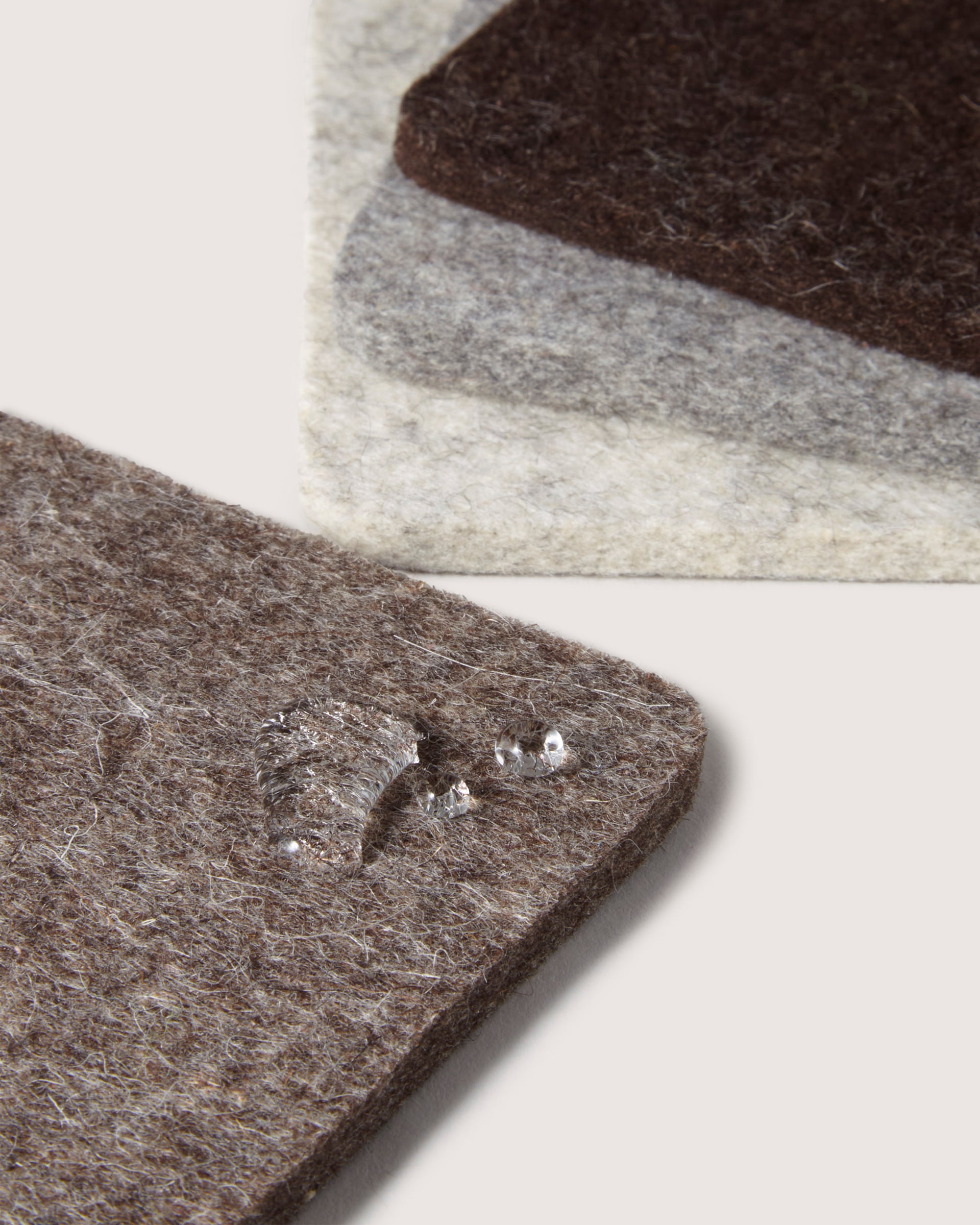 Close-up of three stacked felt fabric squares in brown, gray, and beige hues by graf lantz. Made from Merino wool, the top brown square has a water droplet emphasizing its texture and potential water resistance. Background is neutral off-white.
