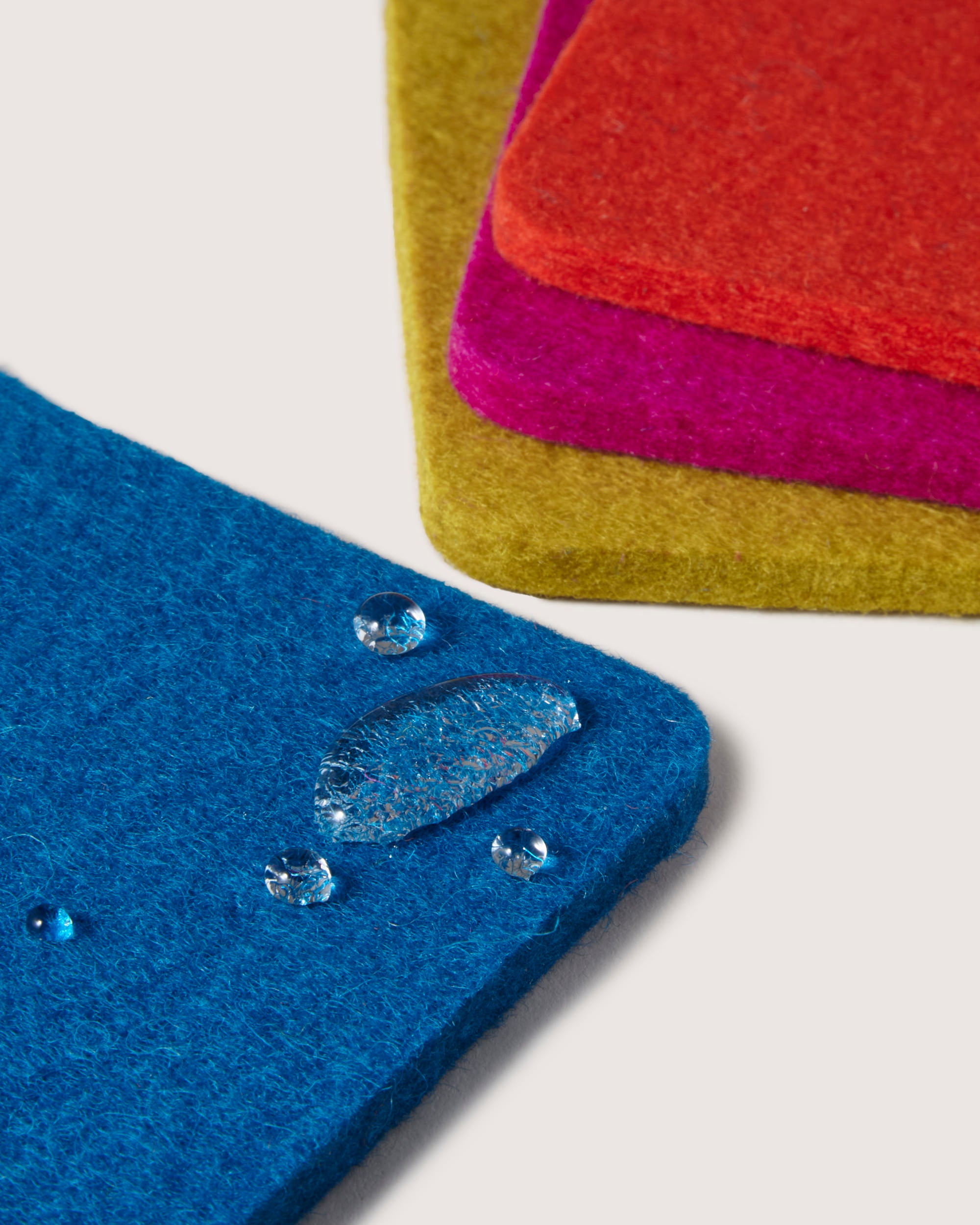 Graf Lantzs colorful Merino wool felt coasters in blue, red, pink, and yellow are stacked on a flat surface. Water droplets glisten on the blue coaster, accentuating its texture and making it a stylish addition to any home décor.