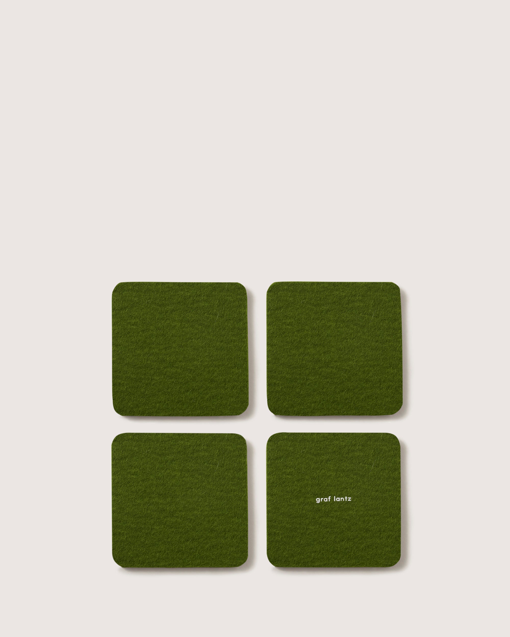 A set of four green square felt coasters, made from luxurious German Merino wool, is laid out in a 2x2 grid on a light surface. One coaster is adorned with graf lantz in white text, highlighting both elegance and practicality.