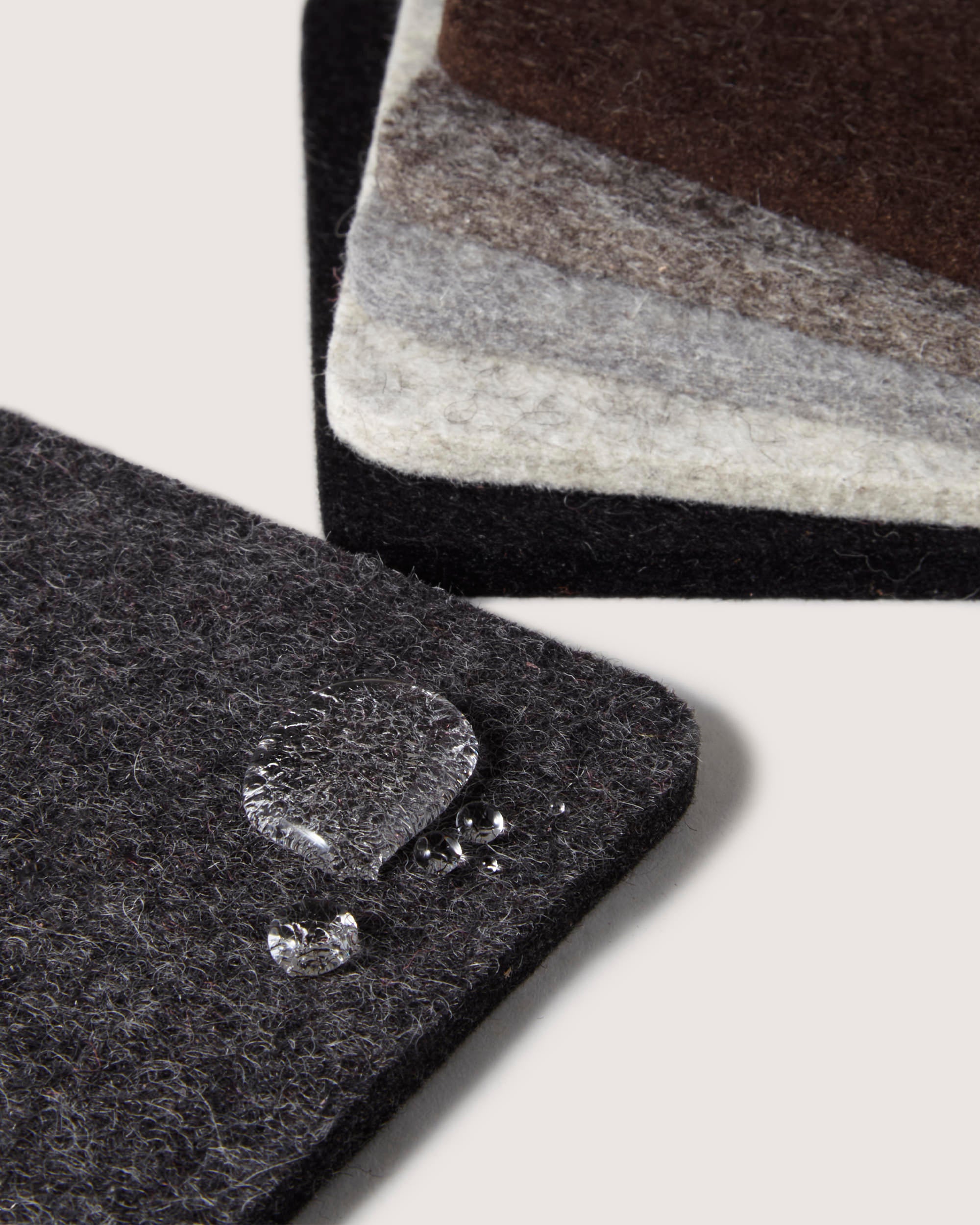 Close-up of four graf lantz sustainable coasters made from German Merino wool in dark gray, light gray, and brown. They are stacked in pairs with water droplets glistening on the top left one, showcasing its textured and water-resistant surface.