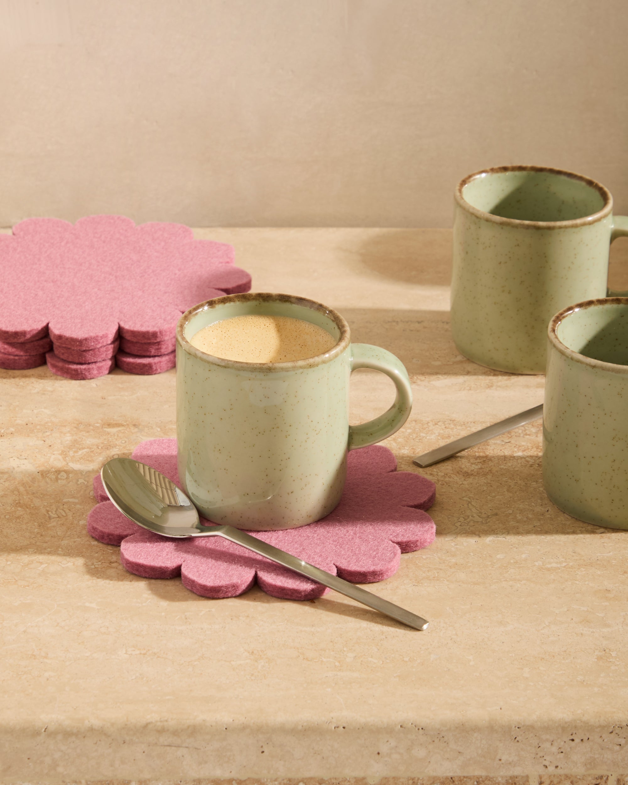 Green ceramic mugs on pink flower-shaped coasters with a spoon on a wooden surface.