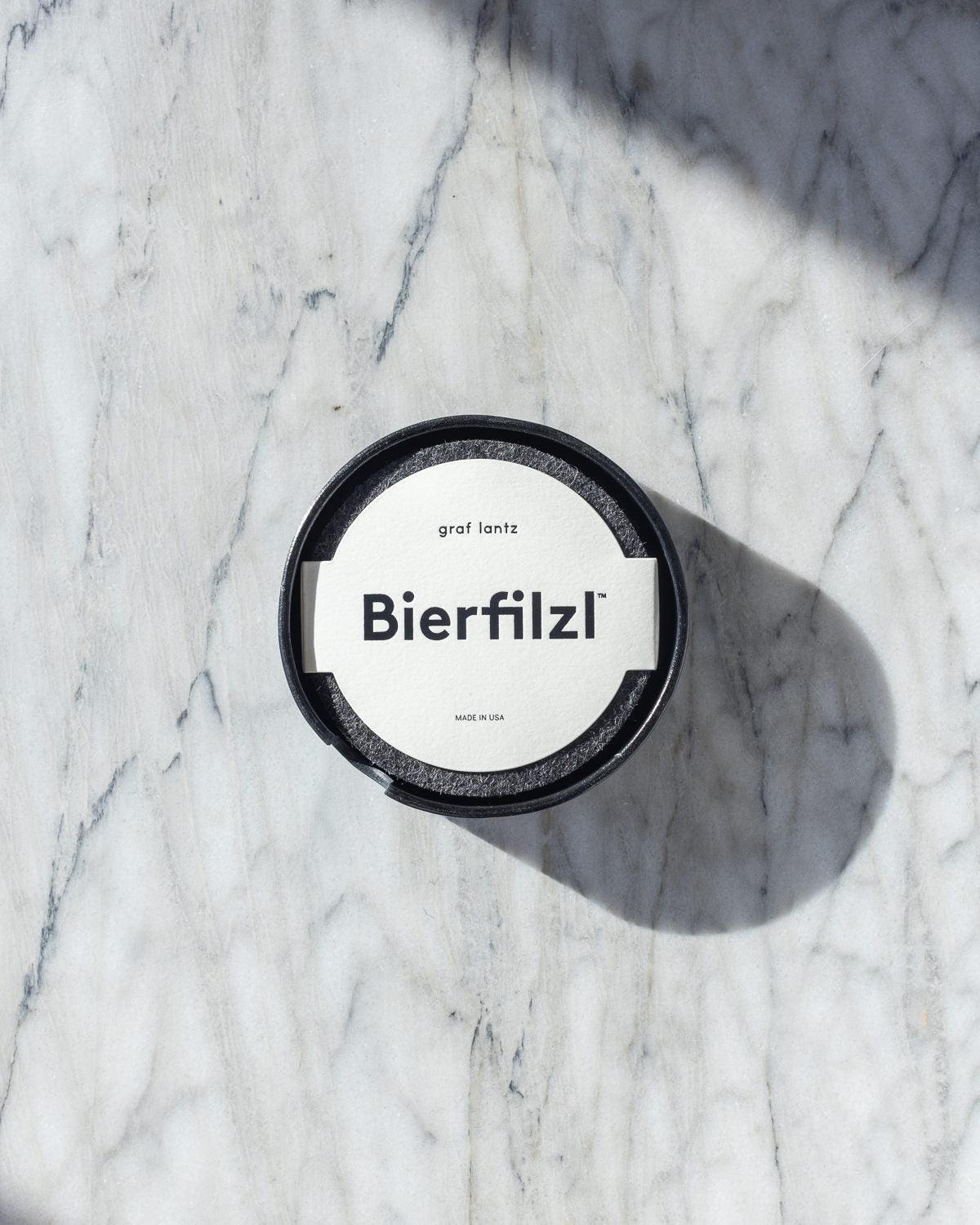A top view of a round black graf lantz container with a white label reading Bierfilzl on light gray marble highlights the elegance of Merino wool craftsmanship. The container casts a shadow to the right, enhancing its sleek design.
