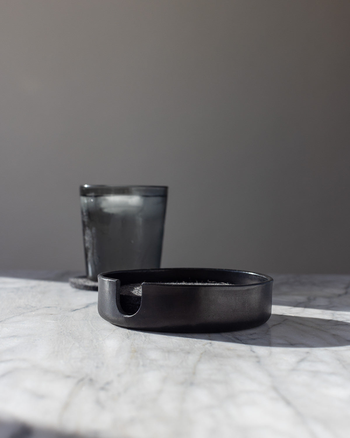 A minimalist scene features a graf lantz dark round container on a marble surface. A frosted glass tumbler with ice stands in the background against a neutral wall, while subtle lighting casts gentle shadows, highlighting its handcrafted elegance.