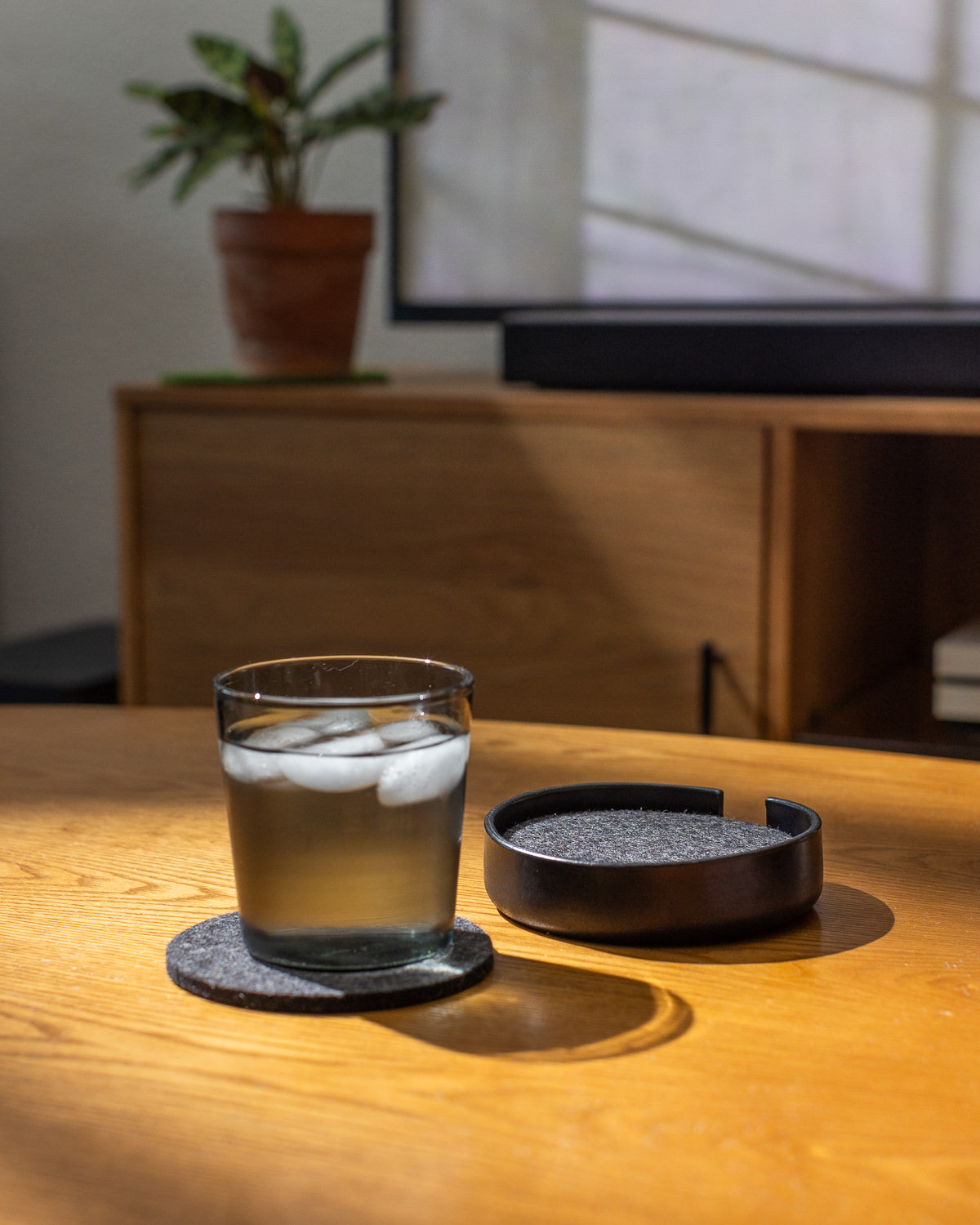 A glass of iced water sits on a graf lantz coaster atop the wooden table, with a handcrafted coaster holder nearby. Soft light casts gentle shadows, while a blurred potted plant in the background adds to the calm and cozy vibes.