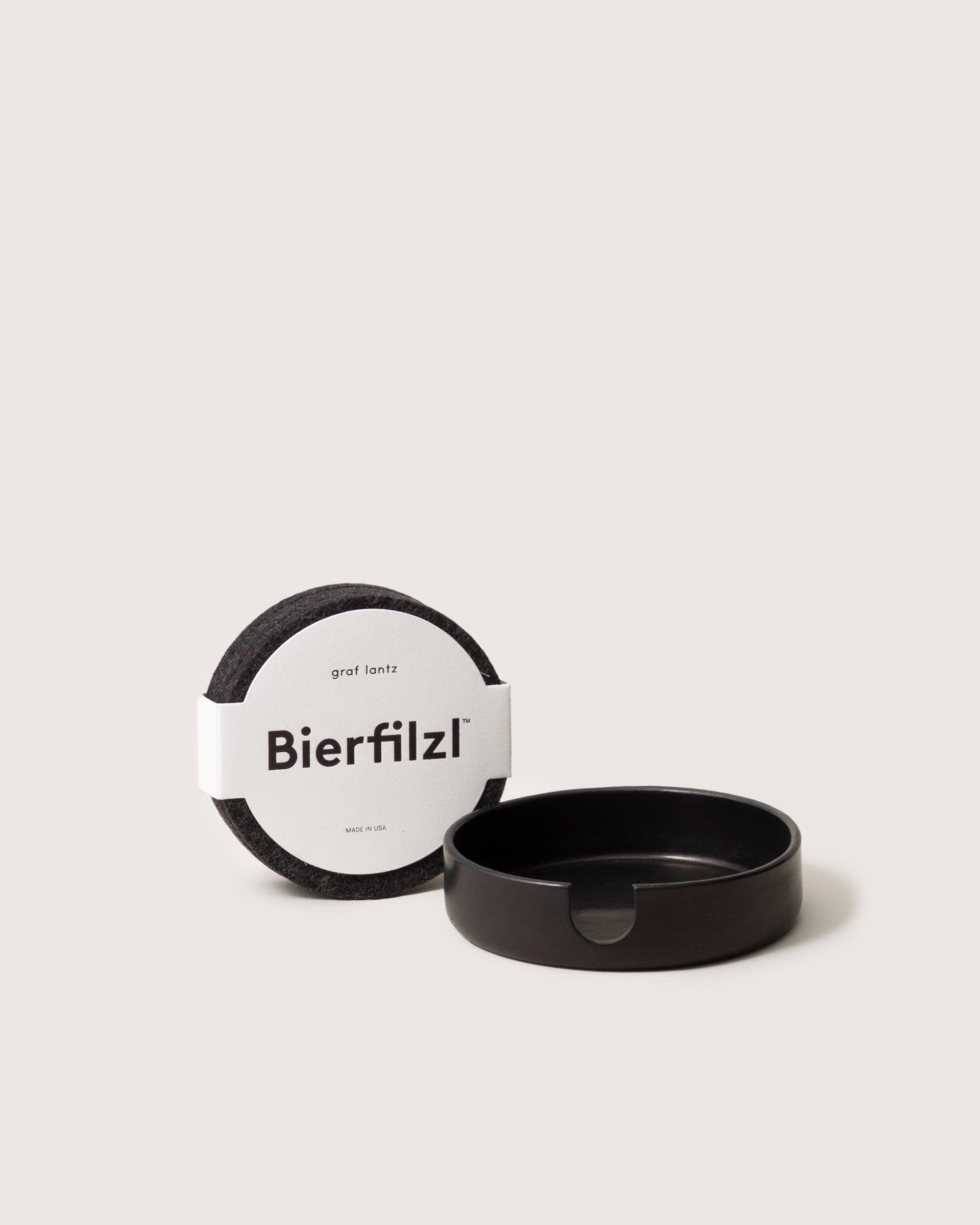 Round gray Bierfilzl coasters crafted from soft Merino wool by graf lantz are paired with a sleek black holder featuring a notch. Set against a light beige background, these handcrafted pieces exude elegance.