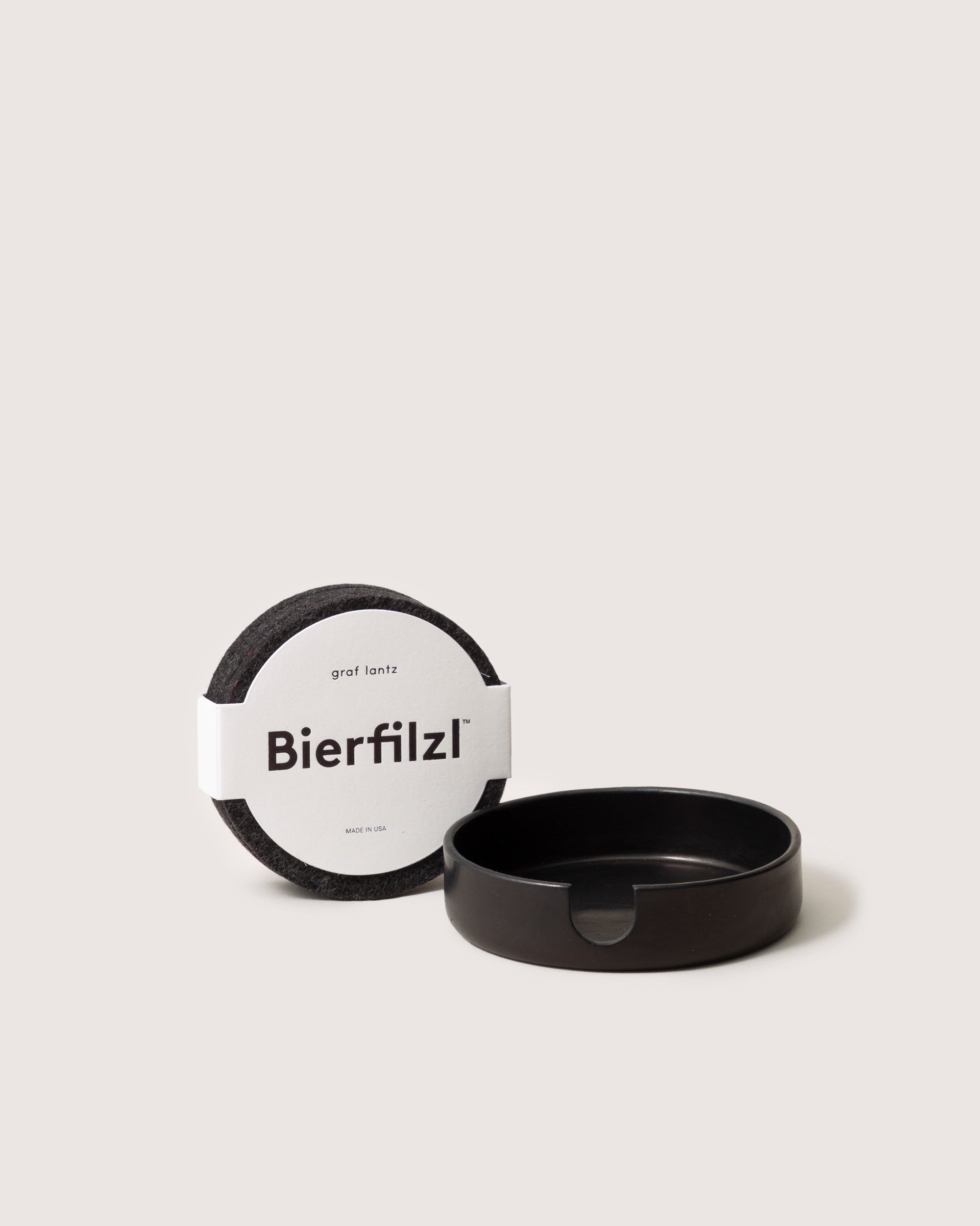 Round gray Bierfilzl coasters crafted from soft Merino wool by graf lantz are paired with a sleek black holder featuring a notch. Set against a light beige background, these handcrafted pieces exude elegance.