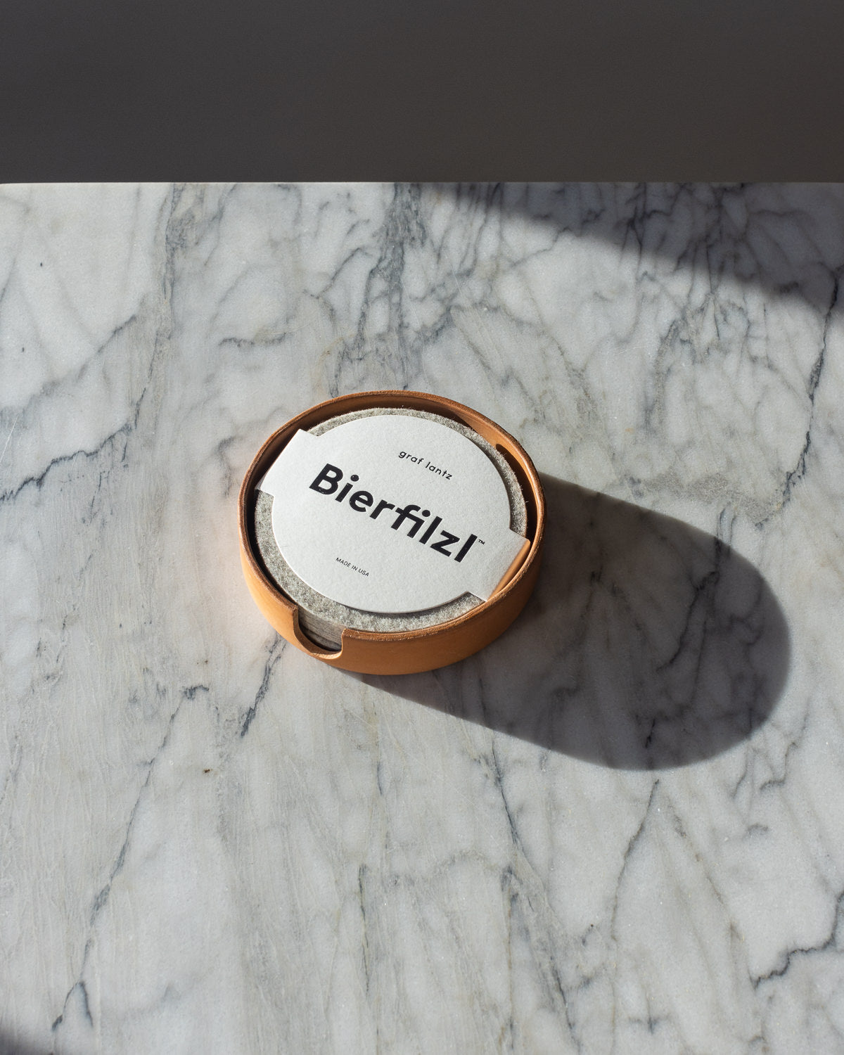 A graf lantz cardboard container with handcrafted Merino wool coasters labeled Bierfilzl sits on a marble surface. The container is partially open, revealing the coasters edge. Shadows on the surface reflect artisanal craftsmanship from Los Angeles.
