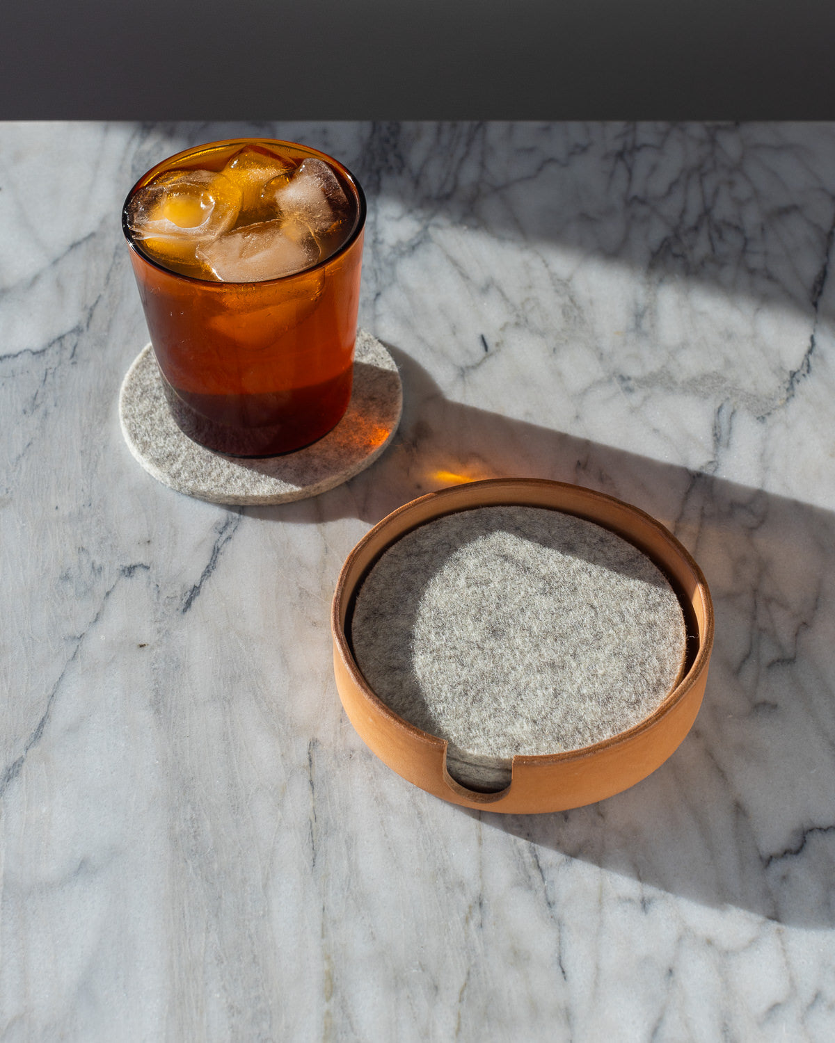 A glass of iced coffee rests on a graf lantz Merino wool felt coaster atop marble, with more coasters stacked in a circular holder. Sunlight casts relaxing shadows.