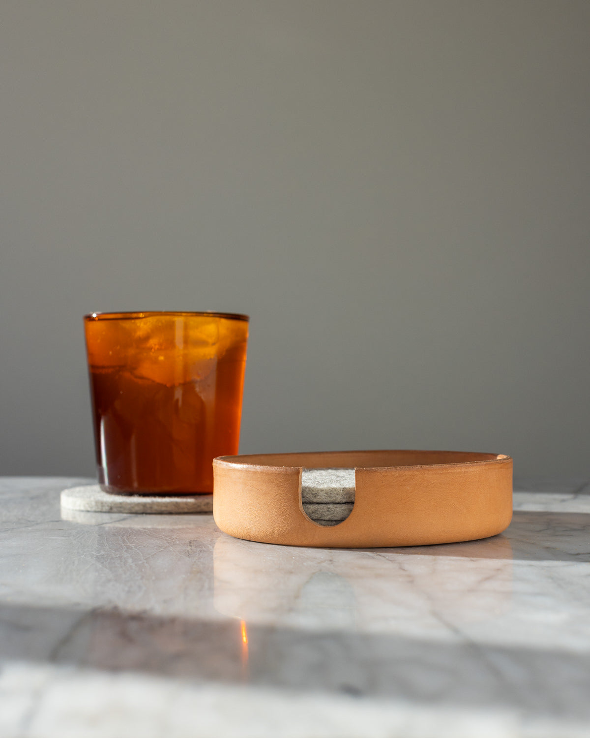 A glass of iced tea rests on a graf lantz Merino wool coaster next to a wooden dish on a marble surface, set against a plain gray background.