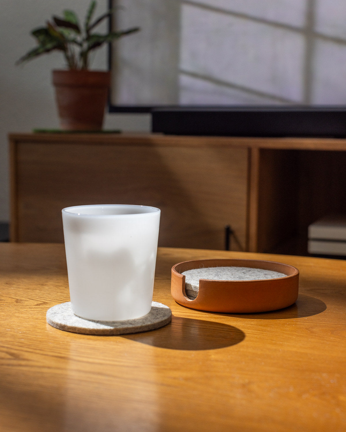 A frosted glass of milk sits on a graf lantz Merino wool coaster atop a wooden table, next to a terracotta-colored coaster holder. A softly lit room reveals a potted plant and part of a wooden cabinet in the background.