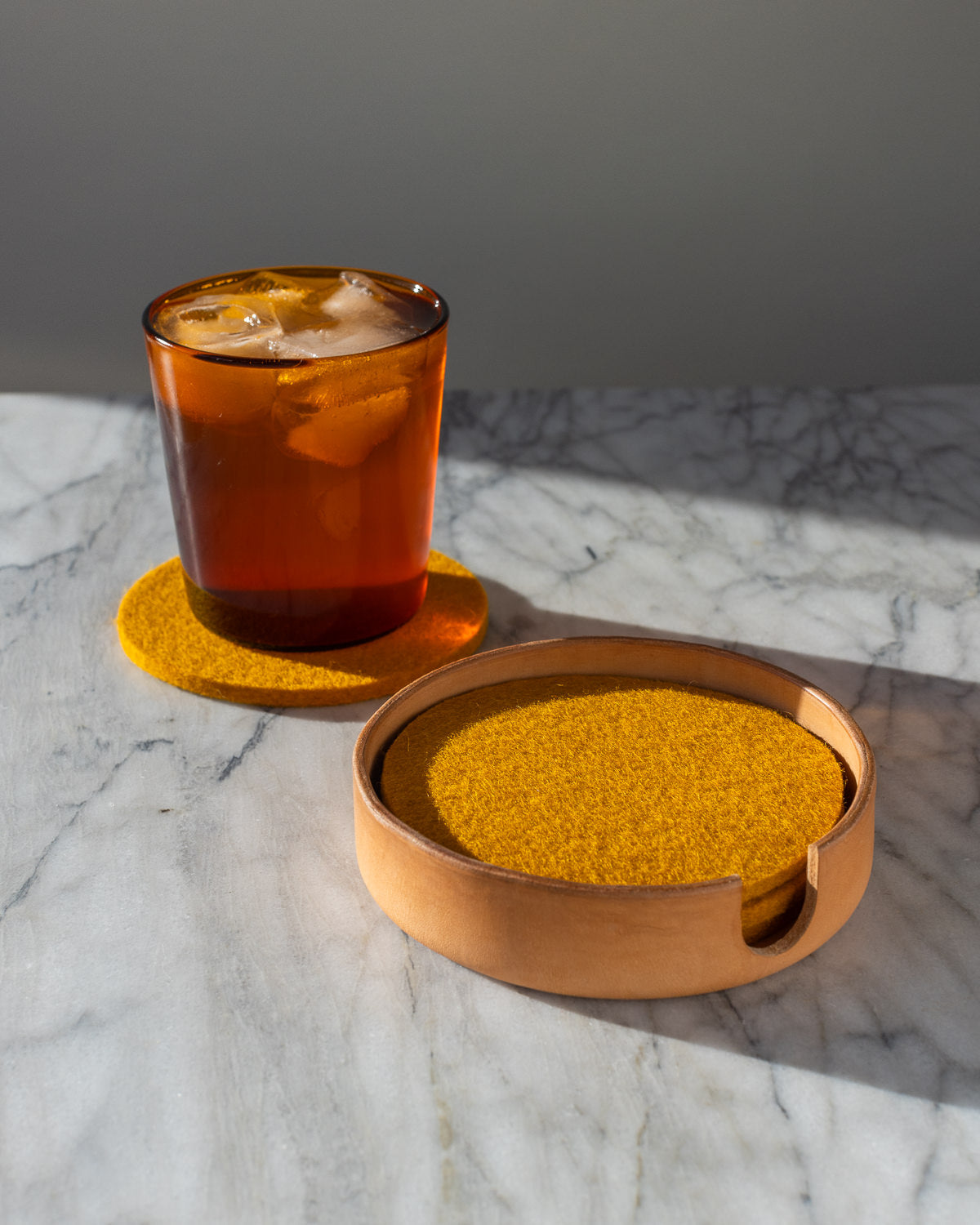A glass filled with an icy beverage sits on a yellow graf lantz felt coaster on a marble surface, with its companion coasters neatly arranged in a wooden holder. Handcrafted coasters gleam under the soft natural light, adding an artisanal touch to the scene.