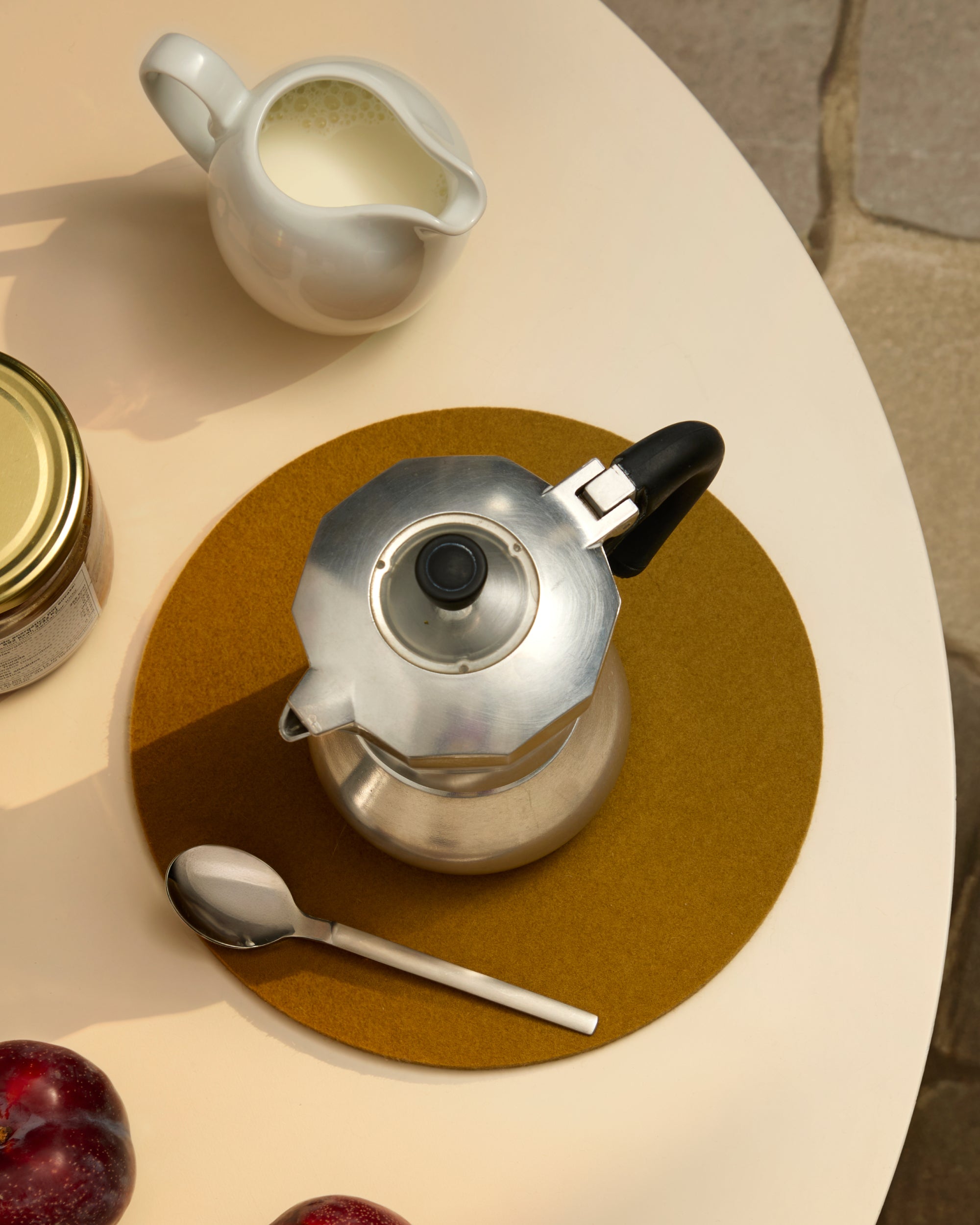 Silver mocapot with black handle on a round Brine trivet, surrounded by a cream pitcher, jar, spoon, and plums on a beige surface.
