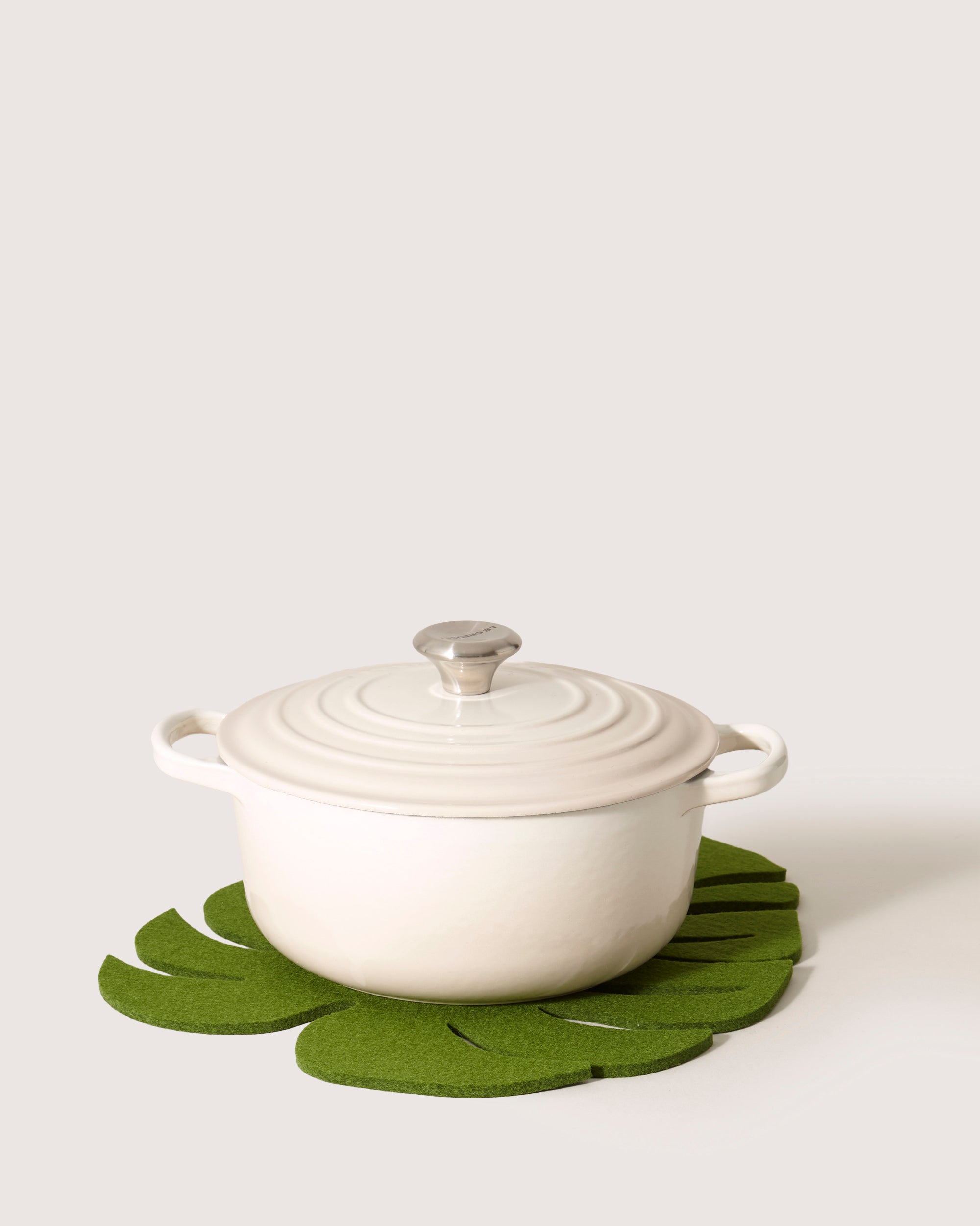 A white cast iron pot with a lid sits on a green, Monstera leaf-shaped trivet by graf lantz, against a plain light background.