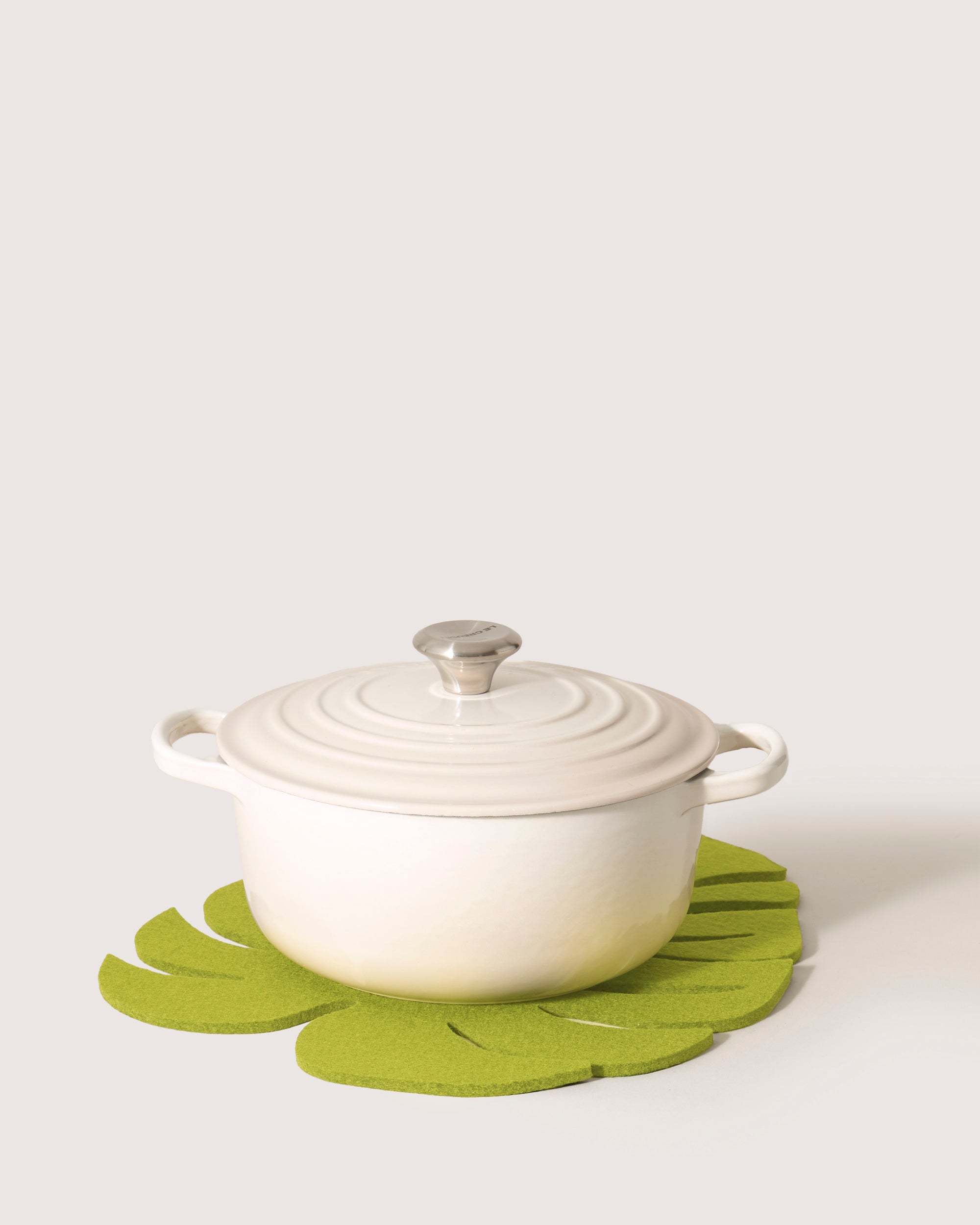 A graf lantz Monstera leaf trivet in green felt holds a white lidded pot with side handles and a metallic knob against a plain, light-colored background.
