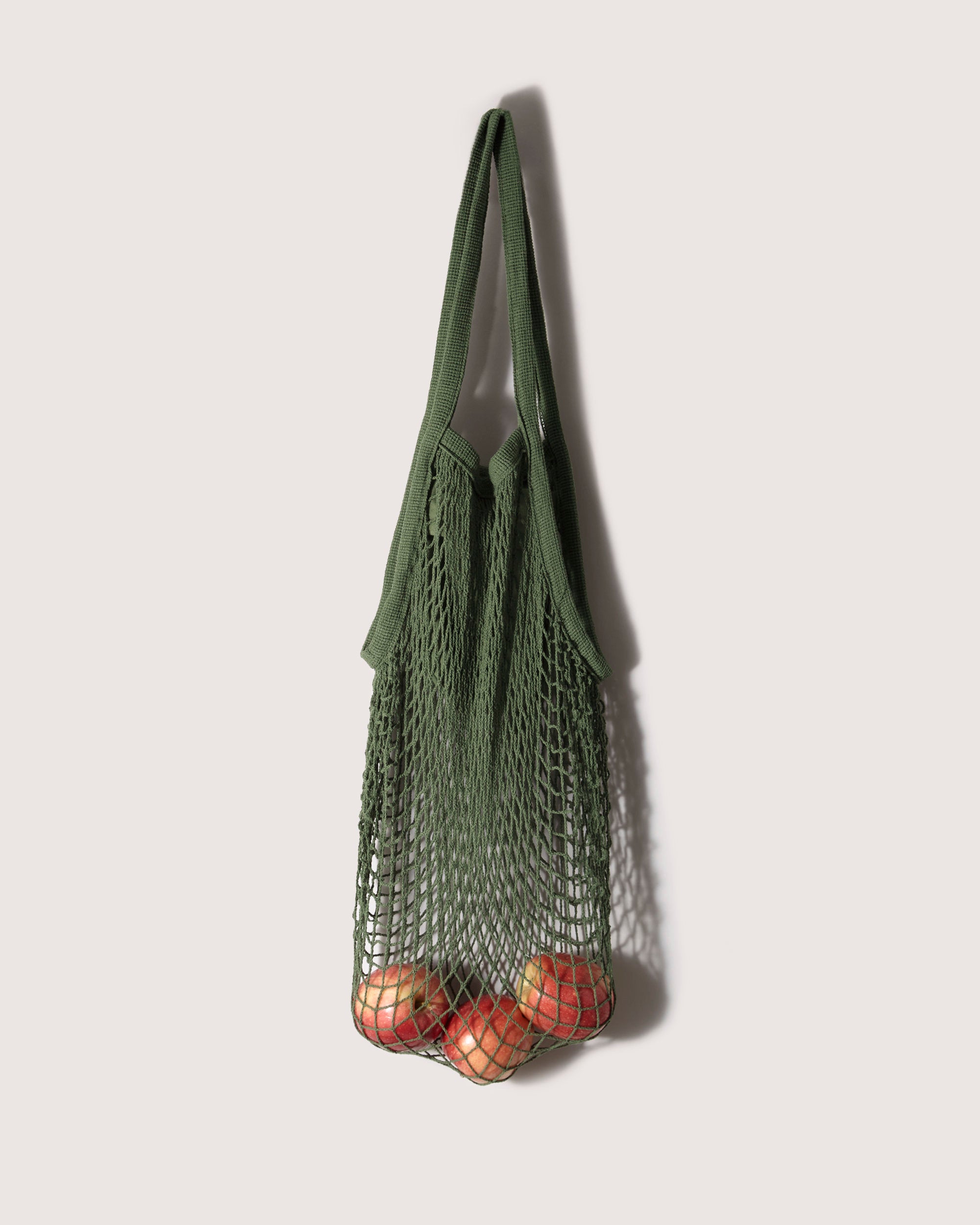 A graf lantz green cotton net tote hangs on the wall, its expandable design perfectly displaying apples at the bottom. The open mesh, similar to a Japanese fish market bag, provides a clear view of the fruit inside.