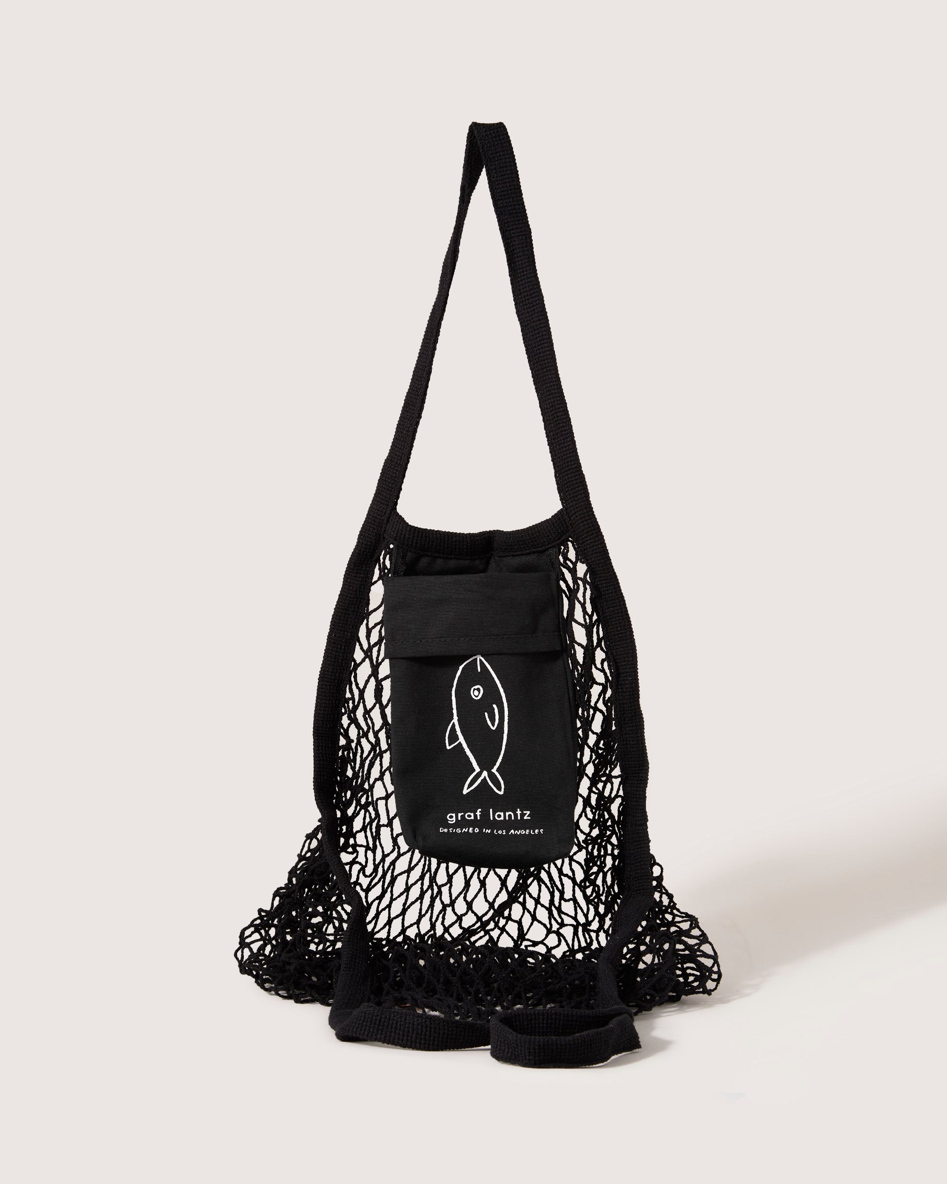 This black cotton netting tote bag by graf lantz features an expandable net design and thick handles. It has a small pocket with a fish graphic, reminiscent of Japanese fish markets, beneath graf lantz on a light gray background.