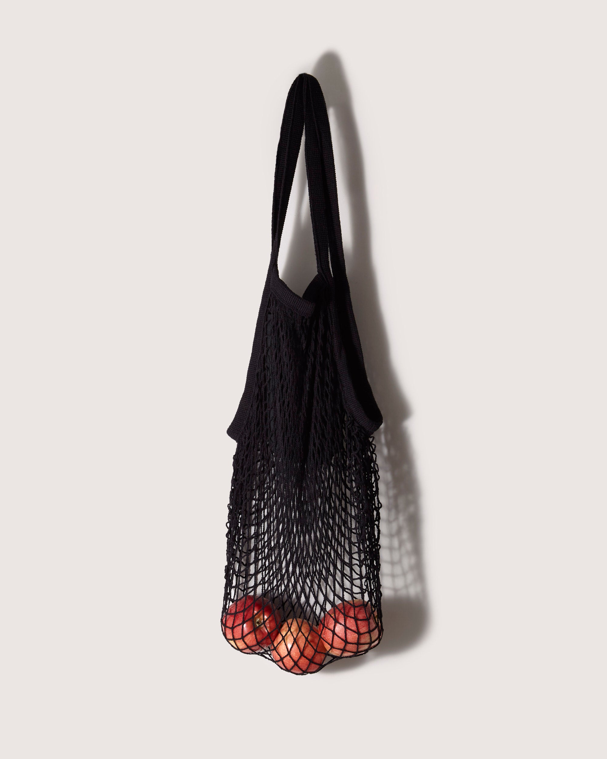 A graf lantz black mesh tote bag with an expandable net design hangs on a light-colored wall, holding a few red apples at the bottom.
