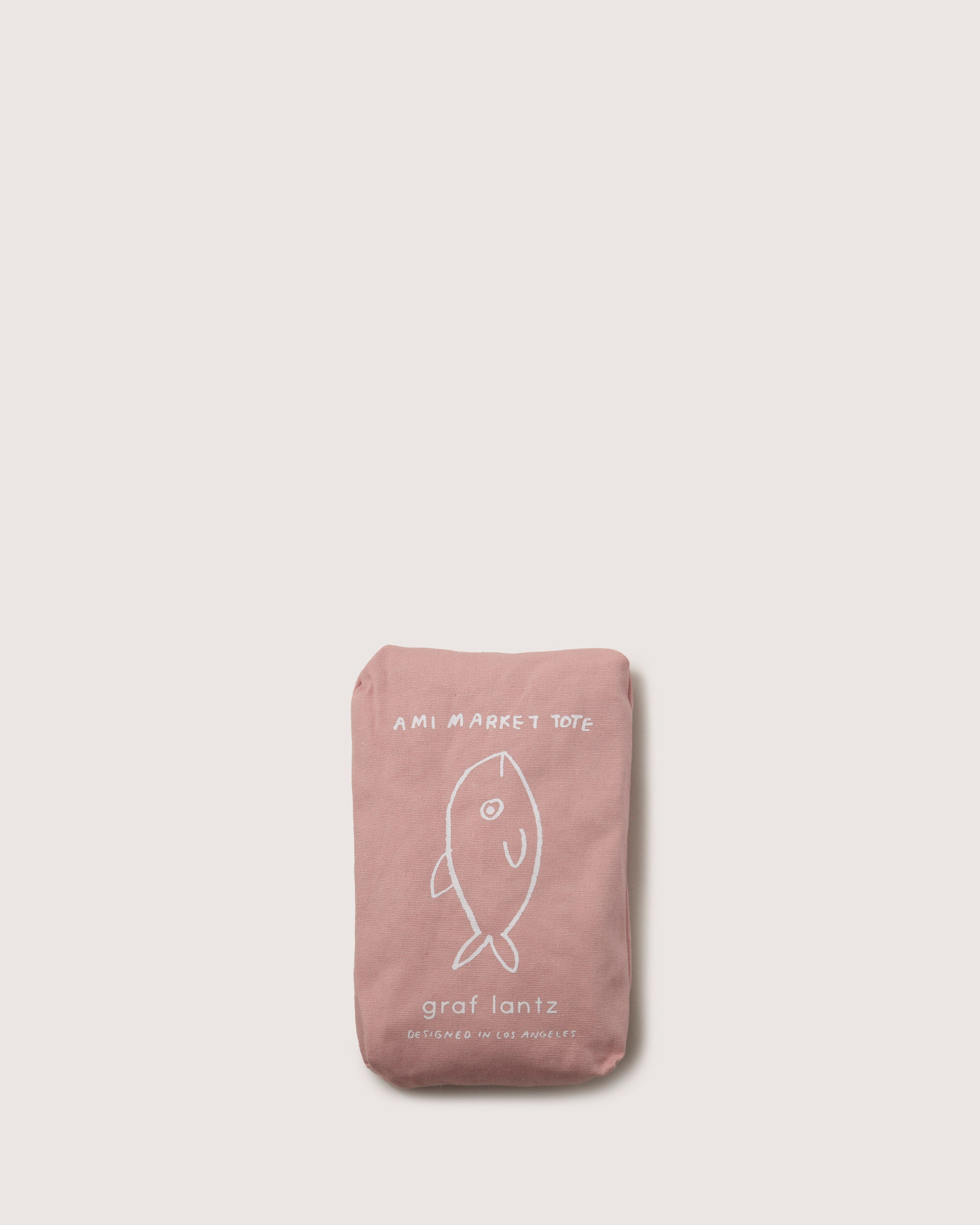 A folded pink tote bag displays a fish line drawing on the front, featuring graf lantz and AMI MARKET TOTE in white lettering. This expandable design blends style and practicality on an off-white background.