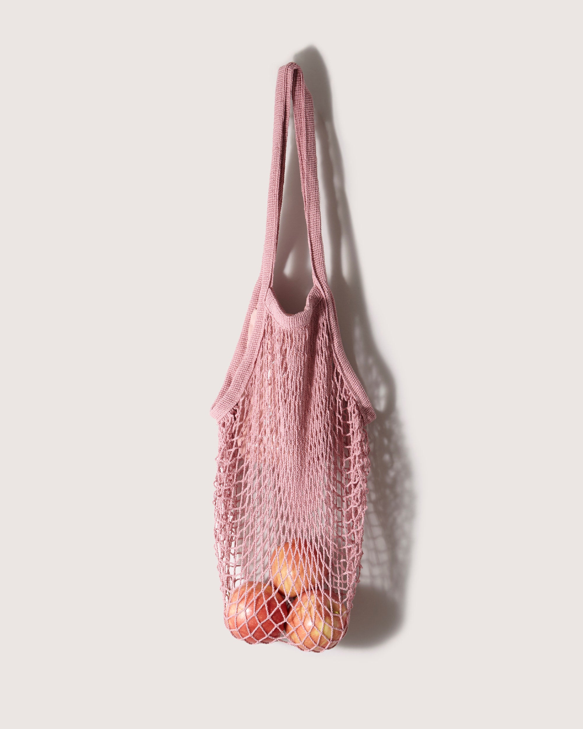 A graf lantz pink cotton netting Net Market Tote, filled with a few oranges, hangs on a wall. Its shadow stretches over the neutral background, highlighting its expandable design.