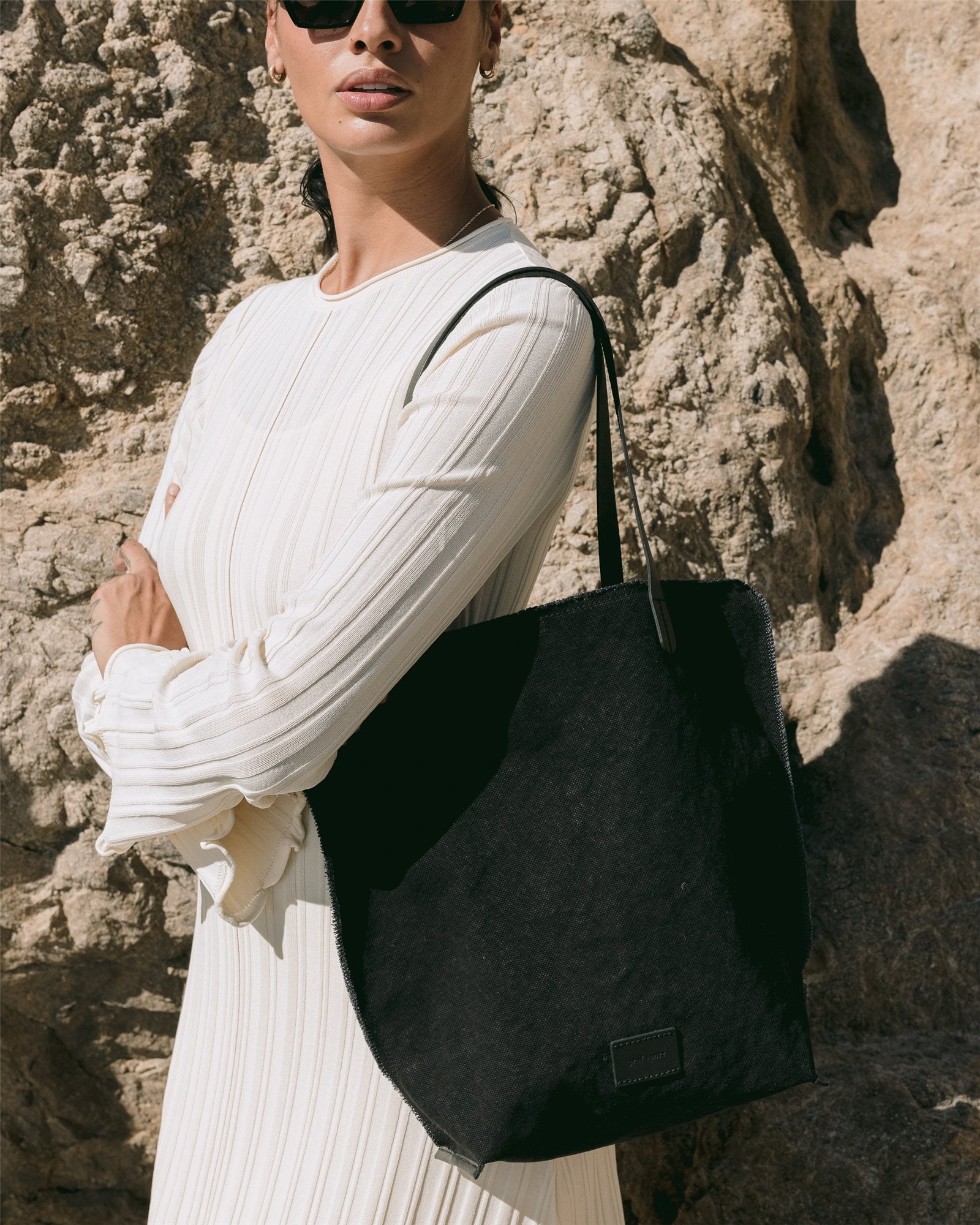A person poses elegantly against a rocky backdrop, wearing sunglasses and a white pleated dress. They hold a stylish Graf Lantz canvas tote with structured leather handles, with their head slightly turned to the side.