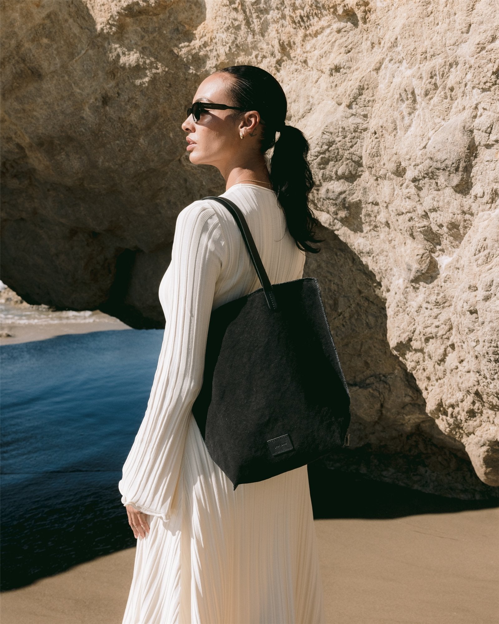 A person stands on a beach by a rocky cliff, wearing sunglasses, a white long-sleeved dress, and carrying a Graf Lantz durable cotton canvas tote with structured leather handles. The ocean and clear sky form a stunning backdrop.