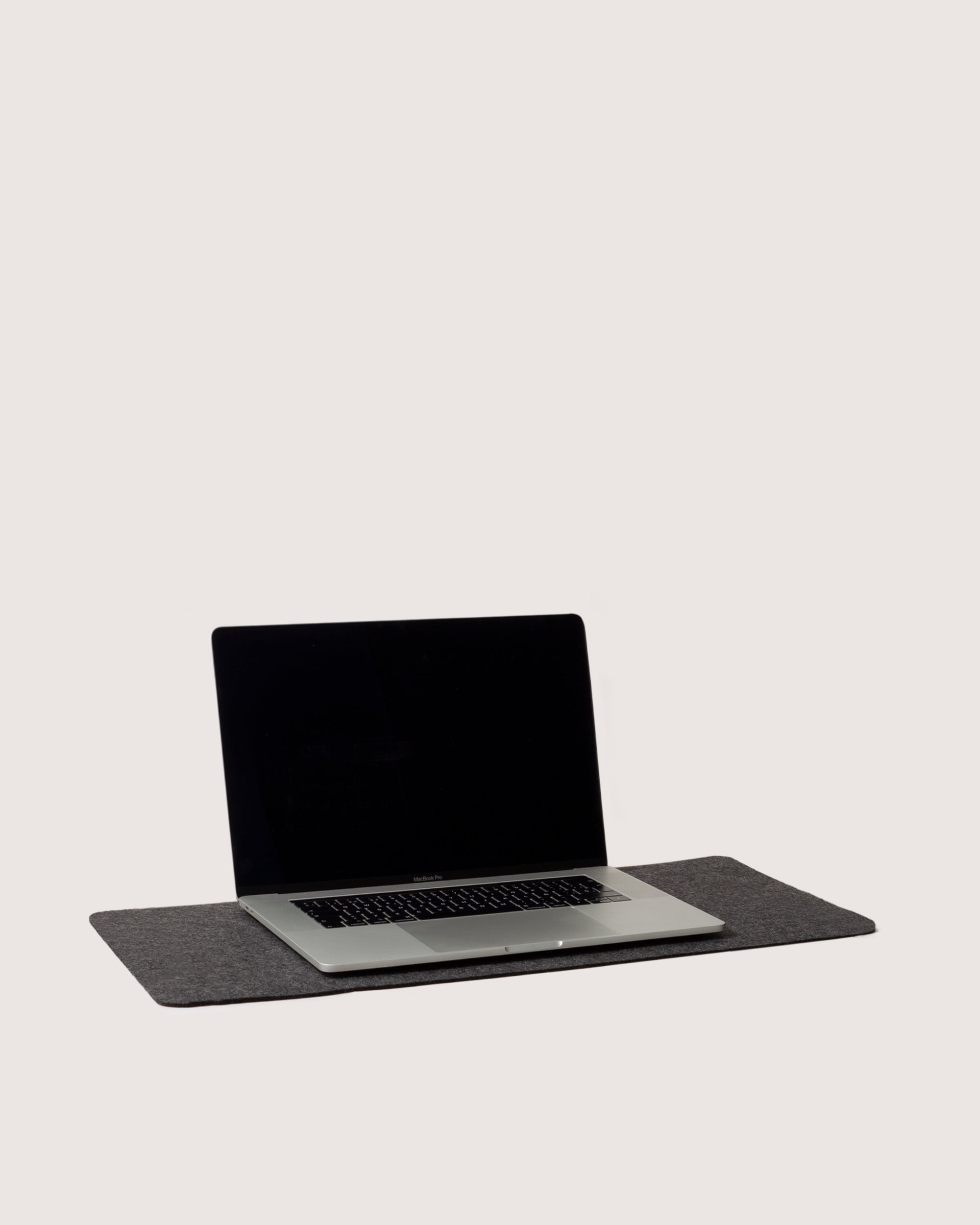 A black-screen laptop rests on a gray graf lantz Merino Wool felt mat, enhancing your workspace aesthetic. Against a plain white background, the open laptop elegantly displays its keyboard with simplicity.