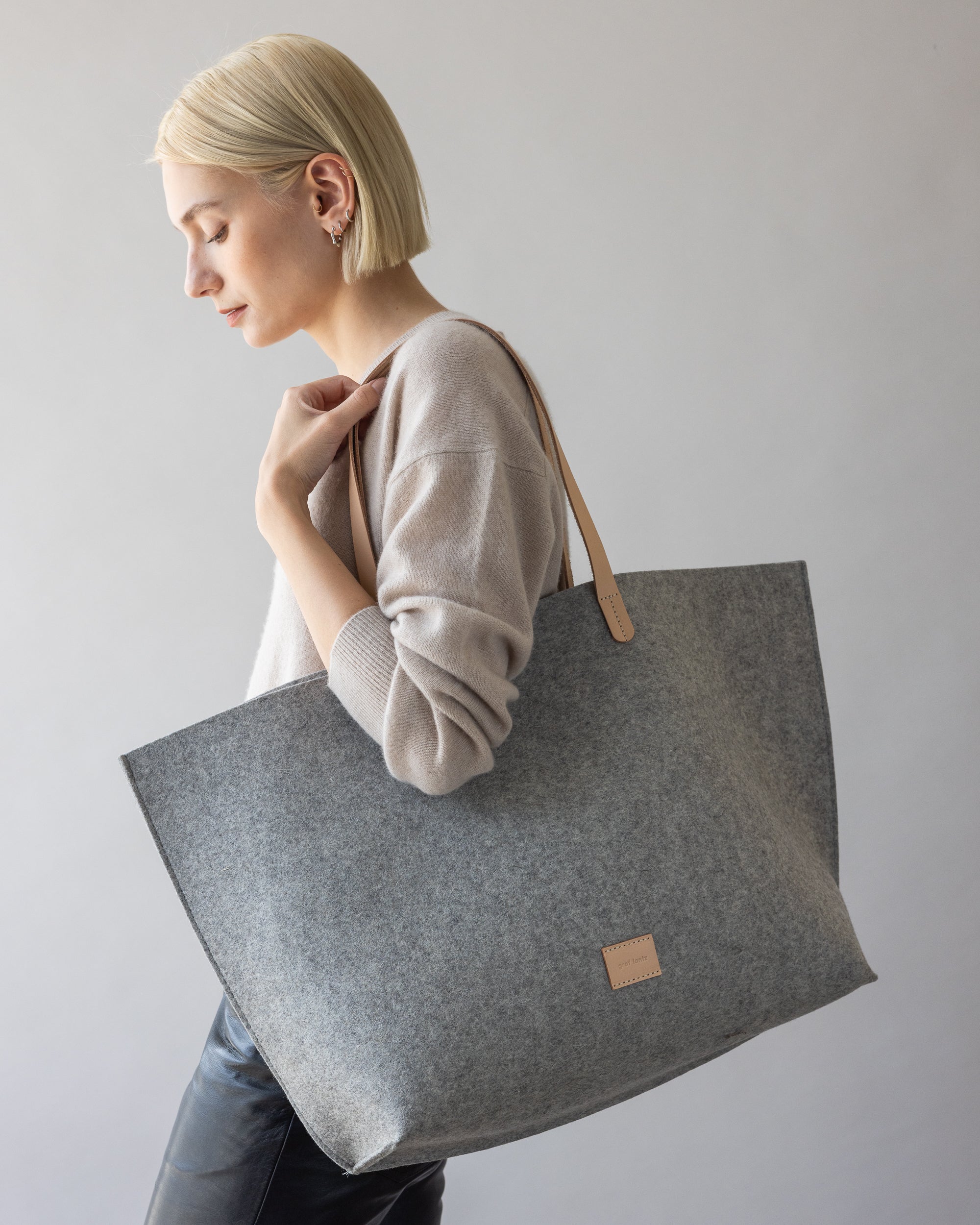 A person with short blonde hair, wearing a beige sweater, carries a large graf lantz gray felt tote bag over their shoulder. Made from Merino wool with leather handles, its minimalist design makes it ideal for beach days.