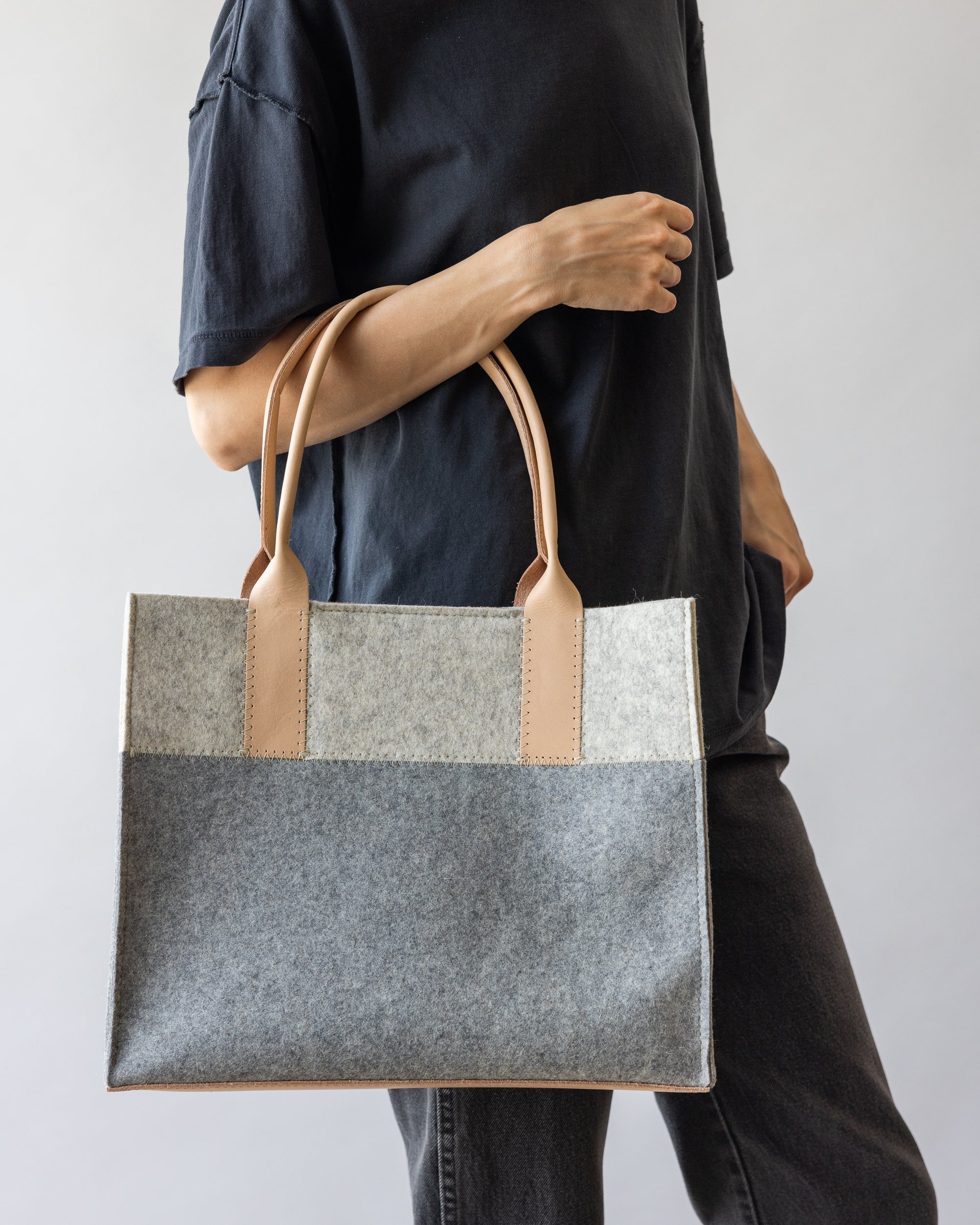 A person in a dark shirt and jeans holds the Jaunt Midi Tote by graf lantz—a versatile mid-size carryall made from two-tone gray felt with tan leather handles. The neutral background complements its stylish and functional design, ideal for any occasion.