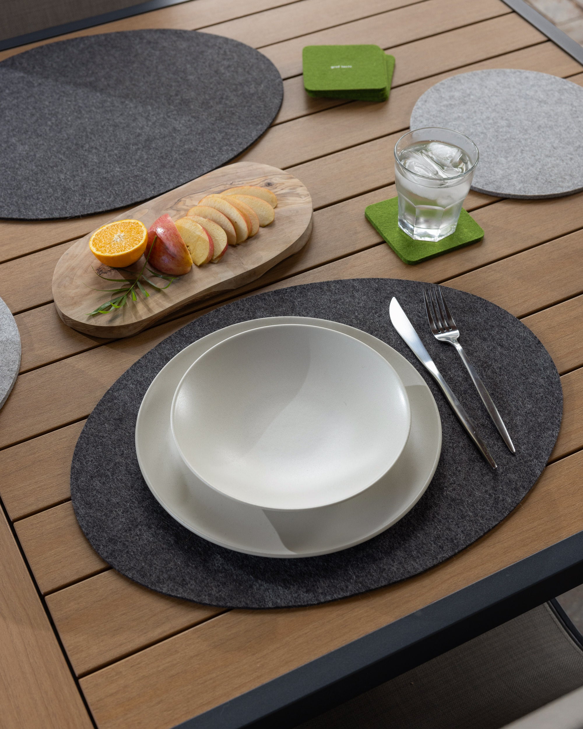A graf lantz Merino wool placemat enhances a wooden table set, featuring a white bowl, plate, silverware, and a glass of water on a green coaster. A wooden tray with sliced apples, orange halves, and rosemary adds natures elegance.