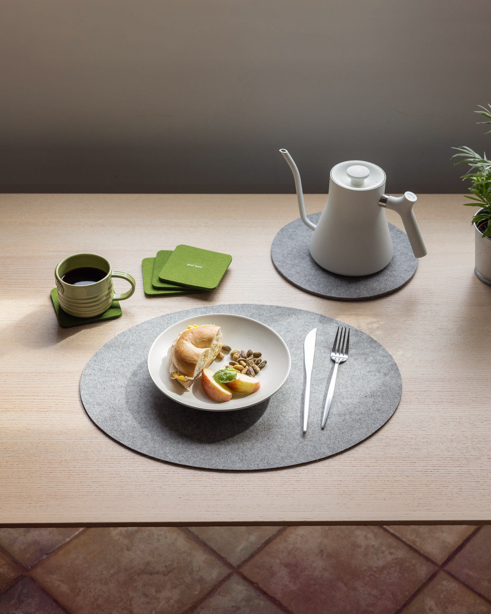 A neatly set table includes a bowl of sliced bread and fruit, with a fork and knife. A green cup sits on a coaster beside a white gooseneck kettle. In the background, theres a potted plant with extra green coasters and elegant graf lantz heat-resistant Merino Wool Placemats.