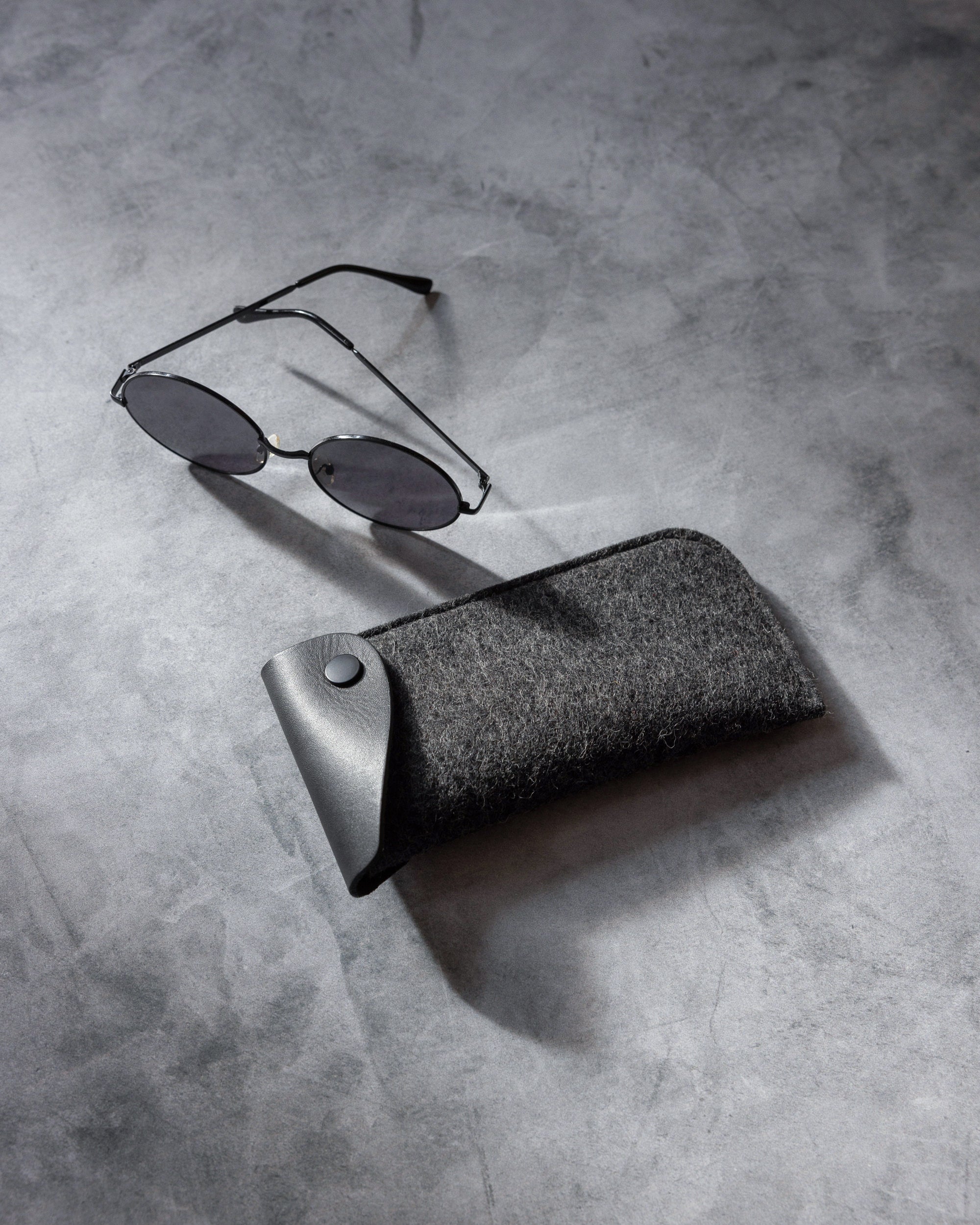 A pair of round sunglasses with thin black frames rests on a gray, textured surface next to a graf lantz Oeko-Tex® Certified felt eyeglass sleeve, featuring a sleek black button closure.