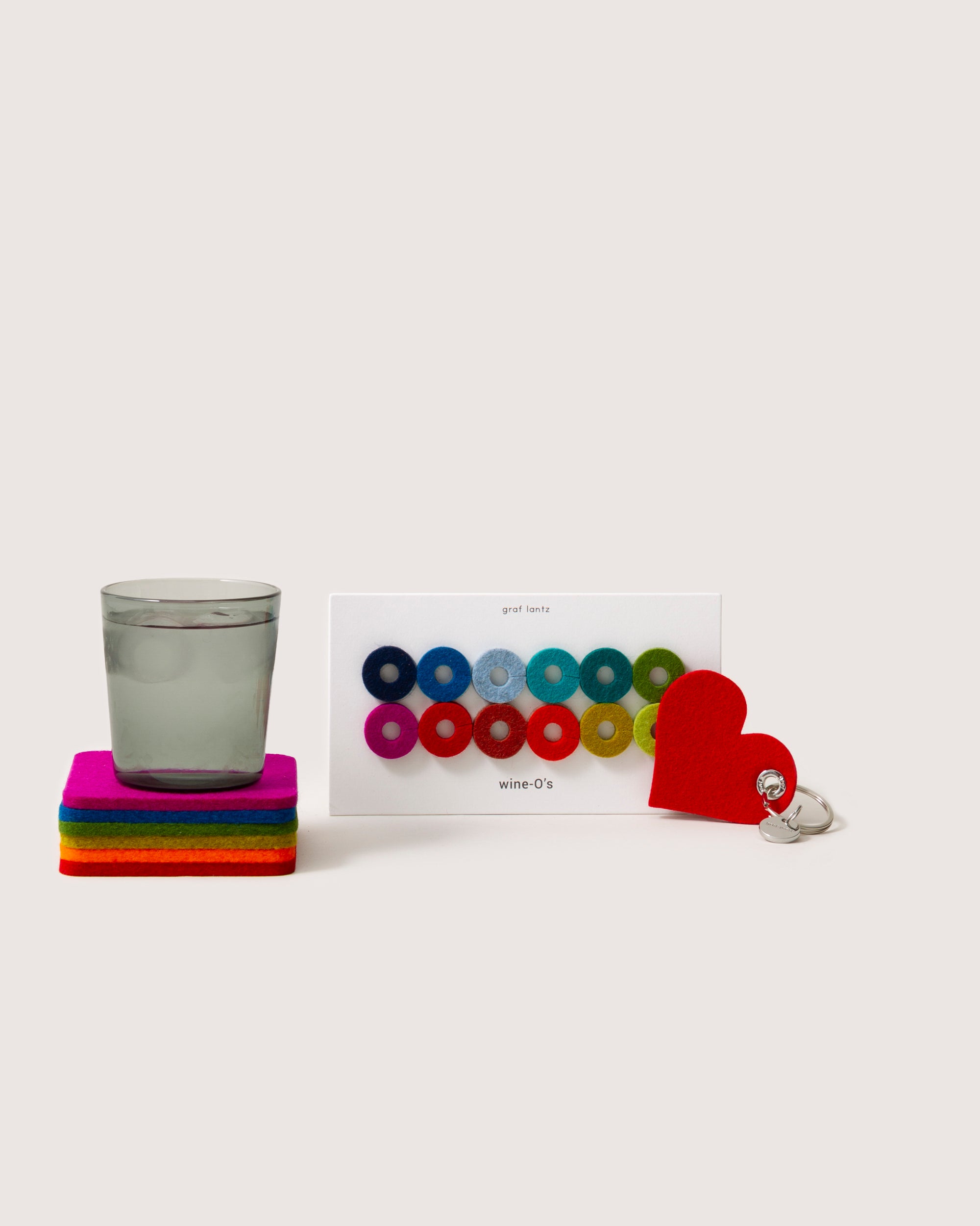 A glass sits on square, colorful graf lantz coasters. Nearby are rainbow wine markers and a heart-shaped red coaster, adding color. A silver wine stopper with a heart top completes the scene against a plain background.